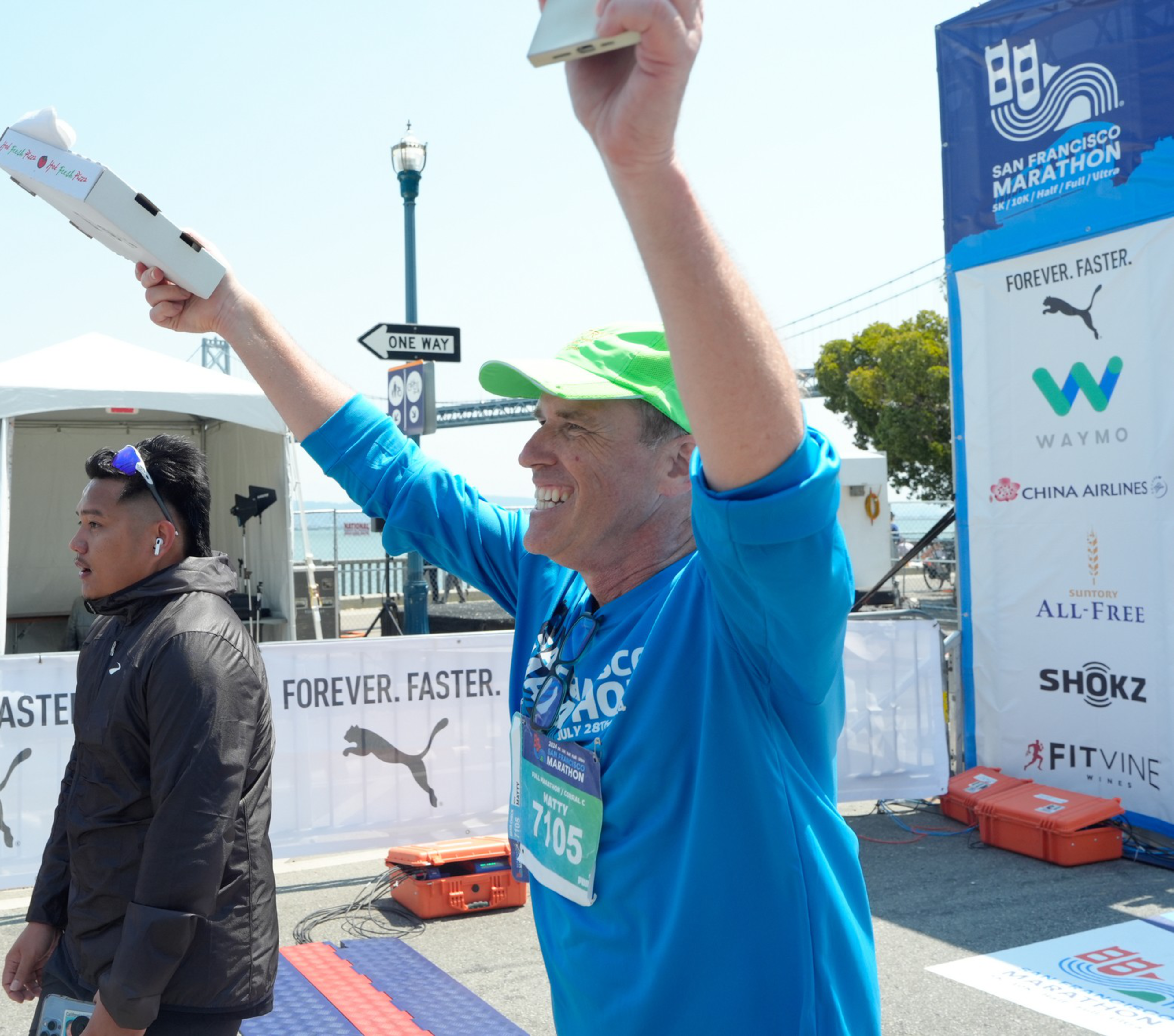 A man in a green cap and blue shirt celebrates at a marathon finish line, holding pizza boxes, while another man in a black jacket walks beside him.
