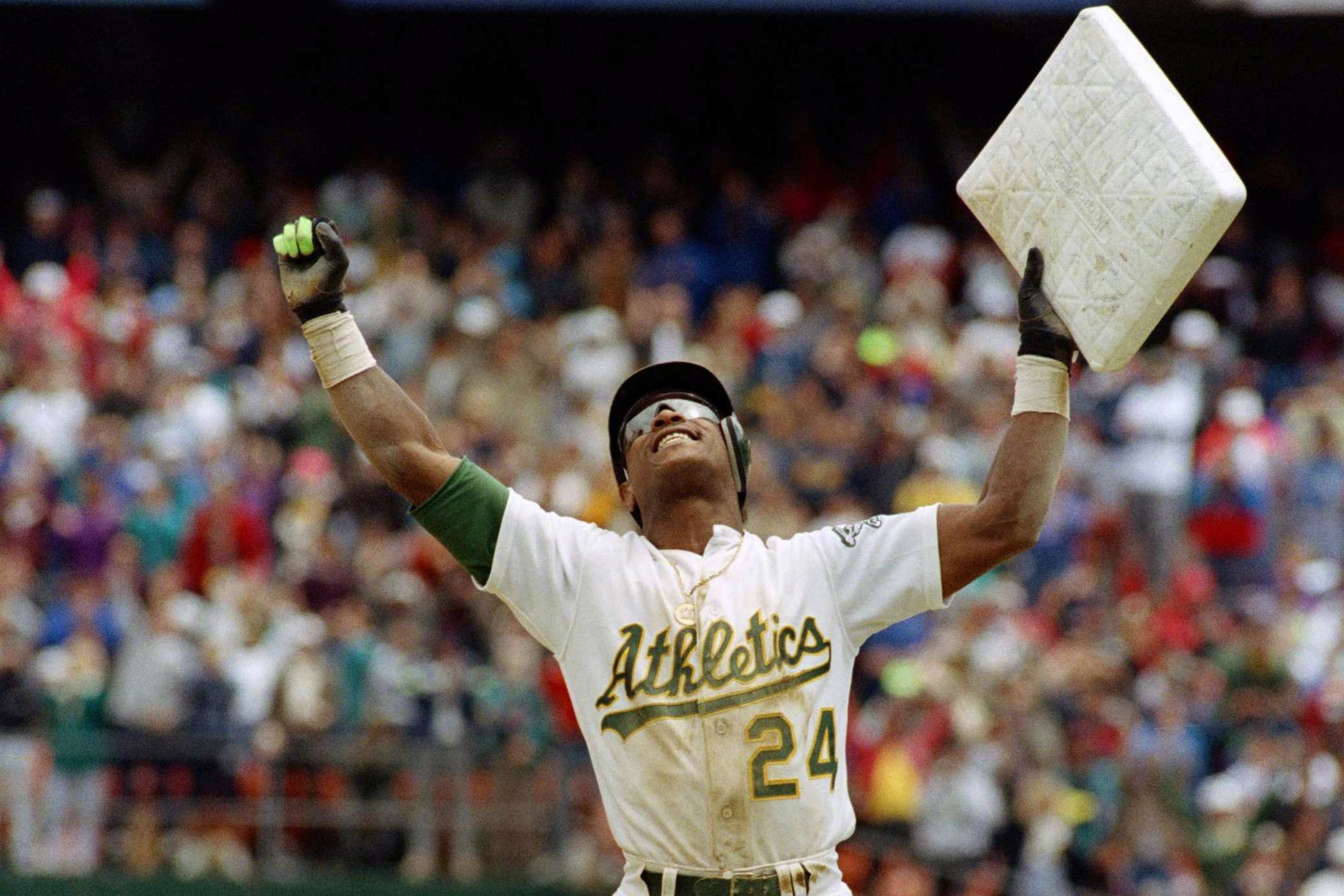 A baseball player in an Athletics uniform, number 24, stands triumphantly holding a base above his head, with a cheering crowd in the background.