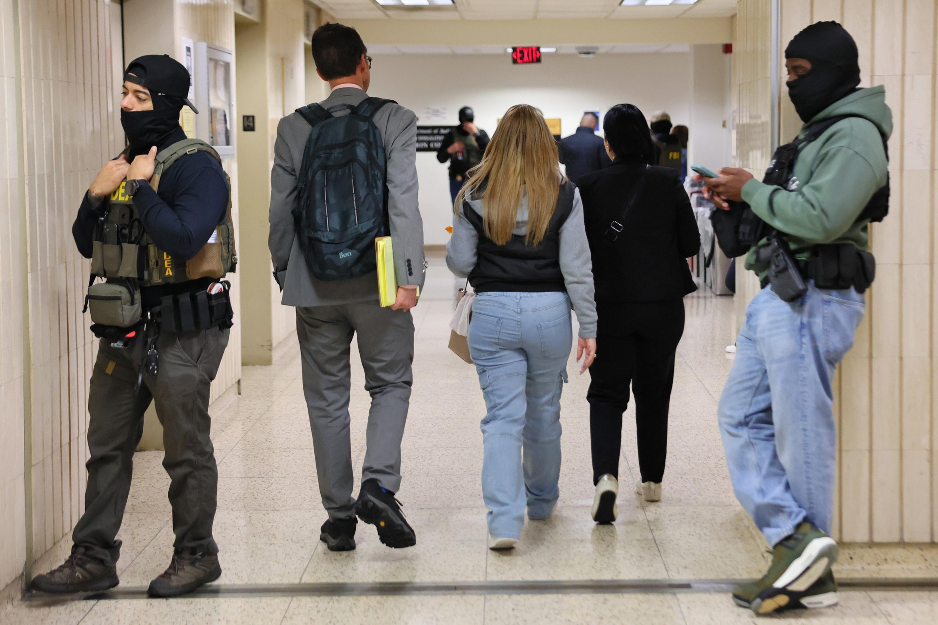 Two masked officers stand guard in a hallway as several people walk away, including a person in a suit and another wearing casual clothing, carrying items.