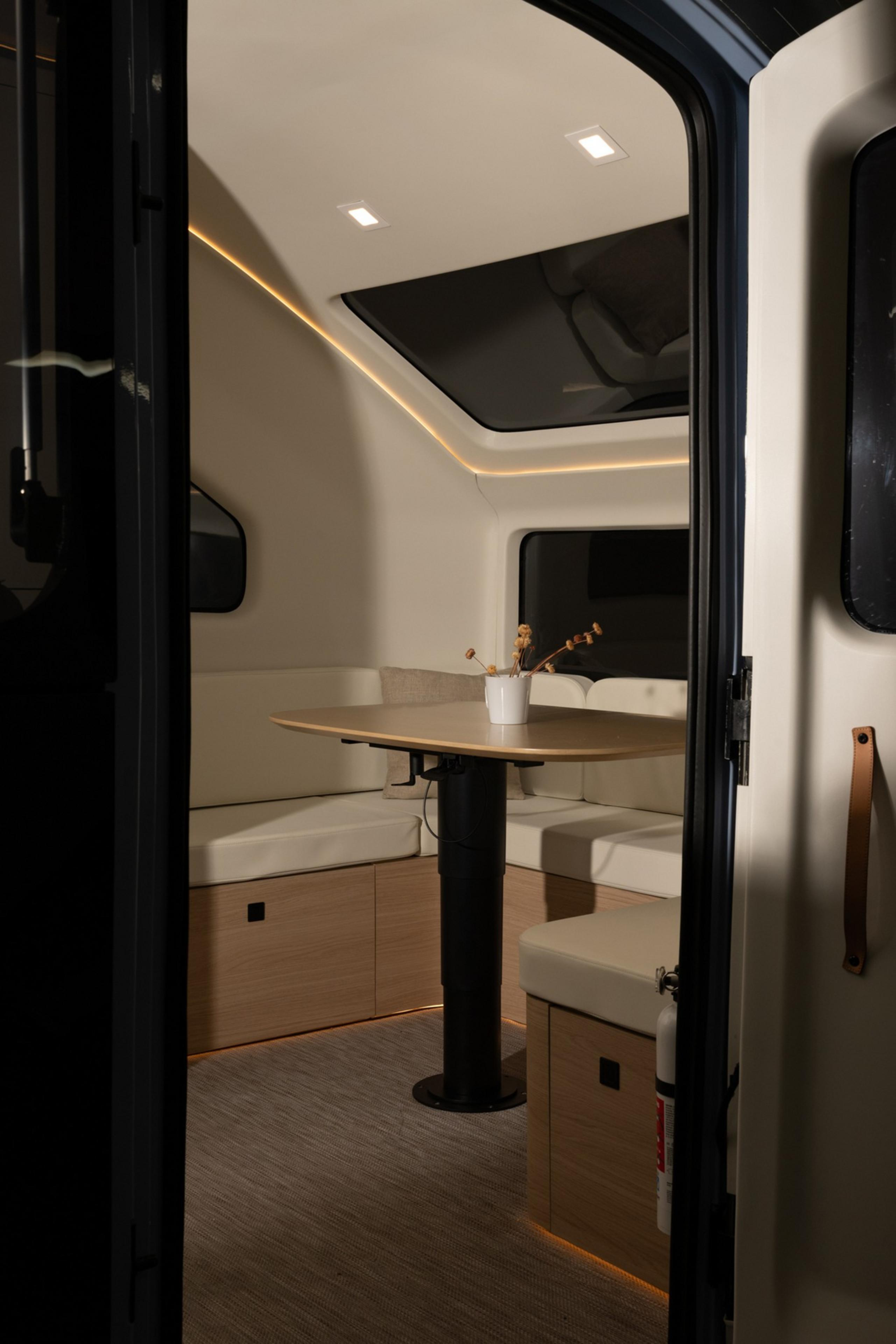 A cozy RV interior with a skylight displays a cushioned seating area around a small table with a potted plant, featuring sleek lighting and wooden accents.
