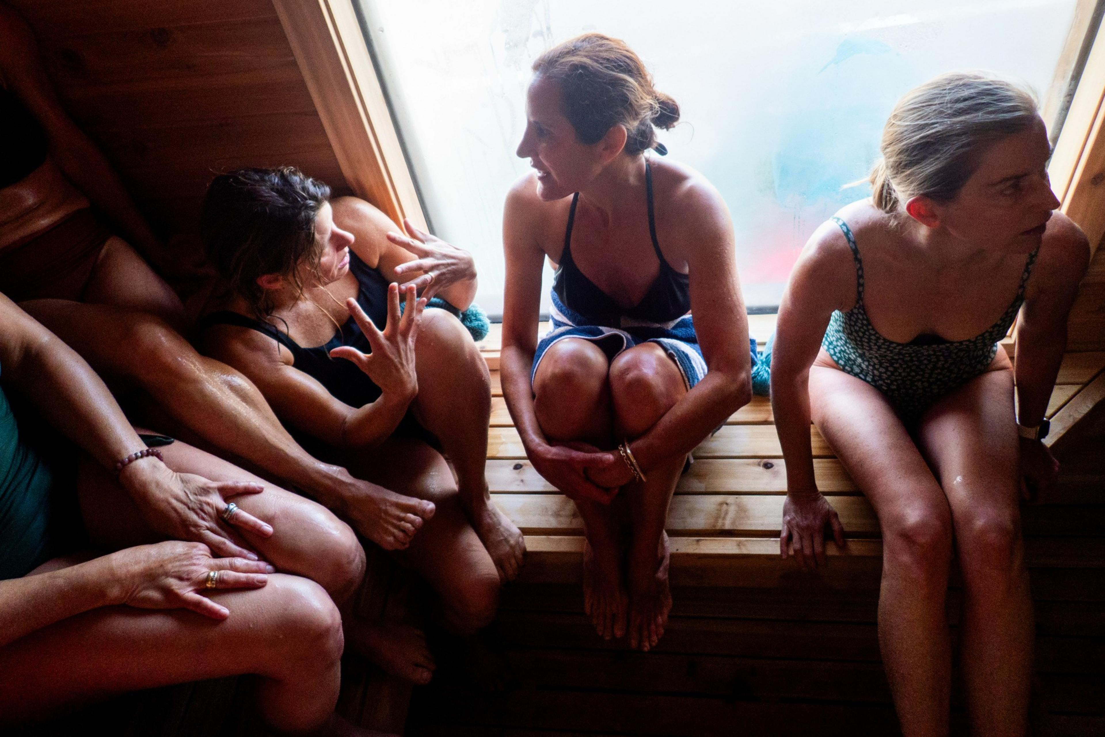 Three women in swimsuits sit closely together on wooden benches in a sauna, engaging in conversation, near a fogged window with light streaming in.