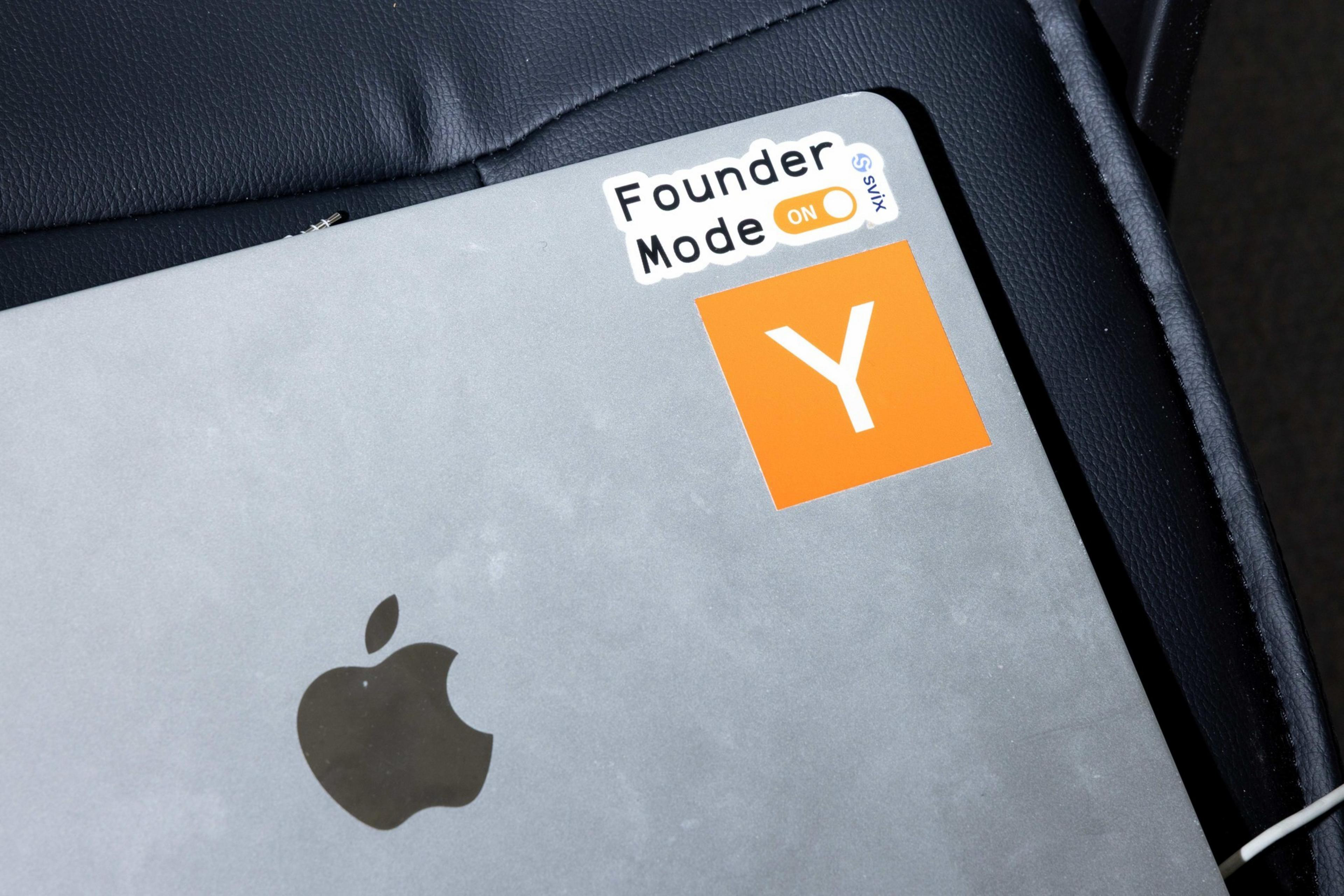 A silver laptop is on a dark surface, featuring an Apple logo and stickers saying &quot;Founder Mode ON&quot; and an orange square with a white &quot;Y.&quot;