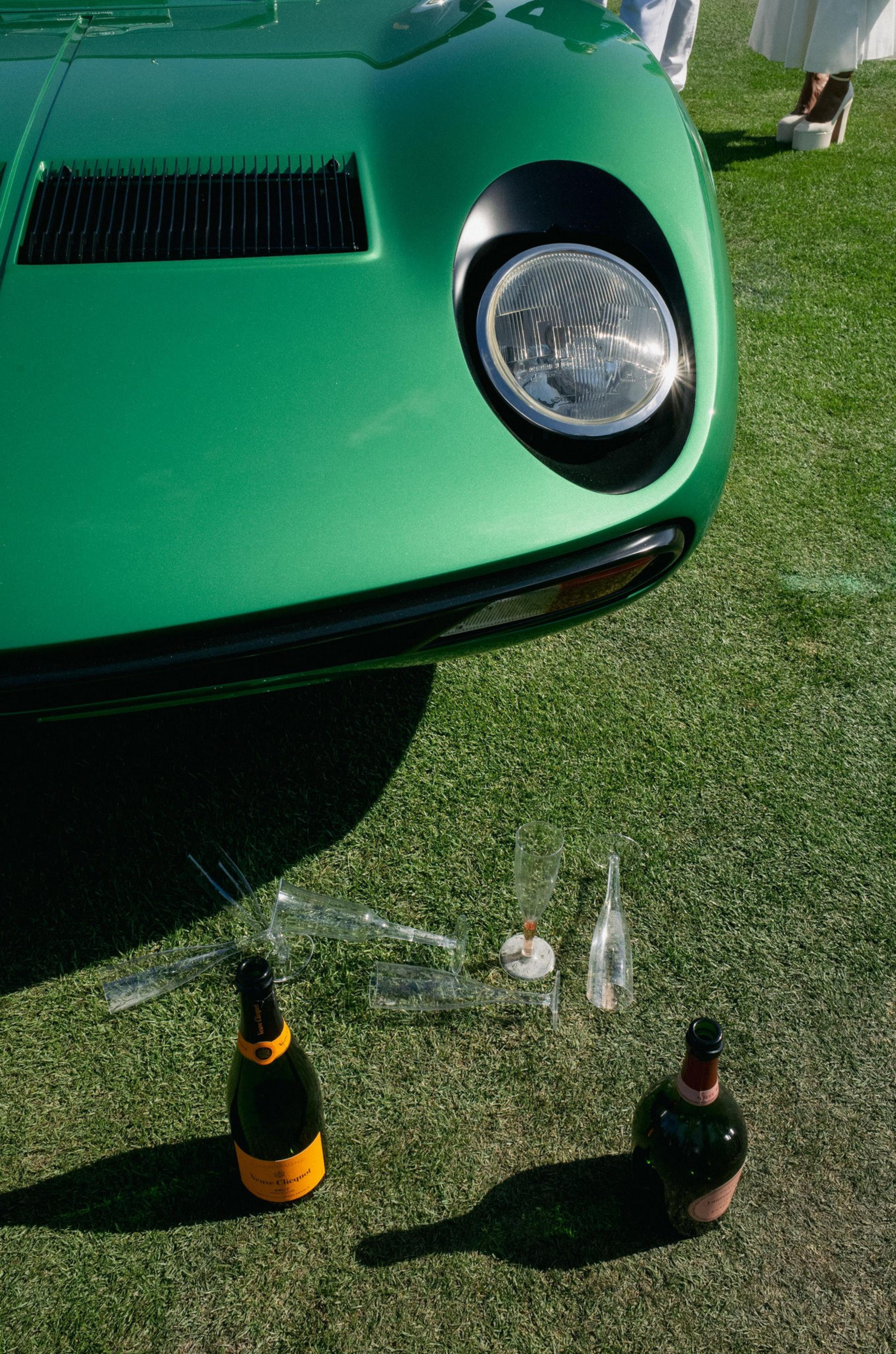 A green classic car's front end with a large round headlight, surrounded by champagne bottles and scattered empty glasses on green grass.
