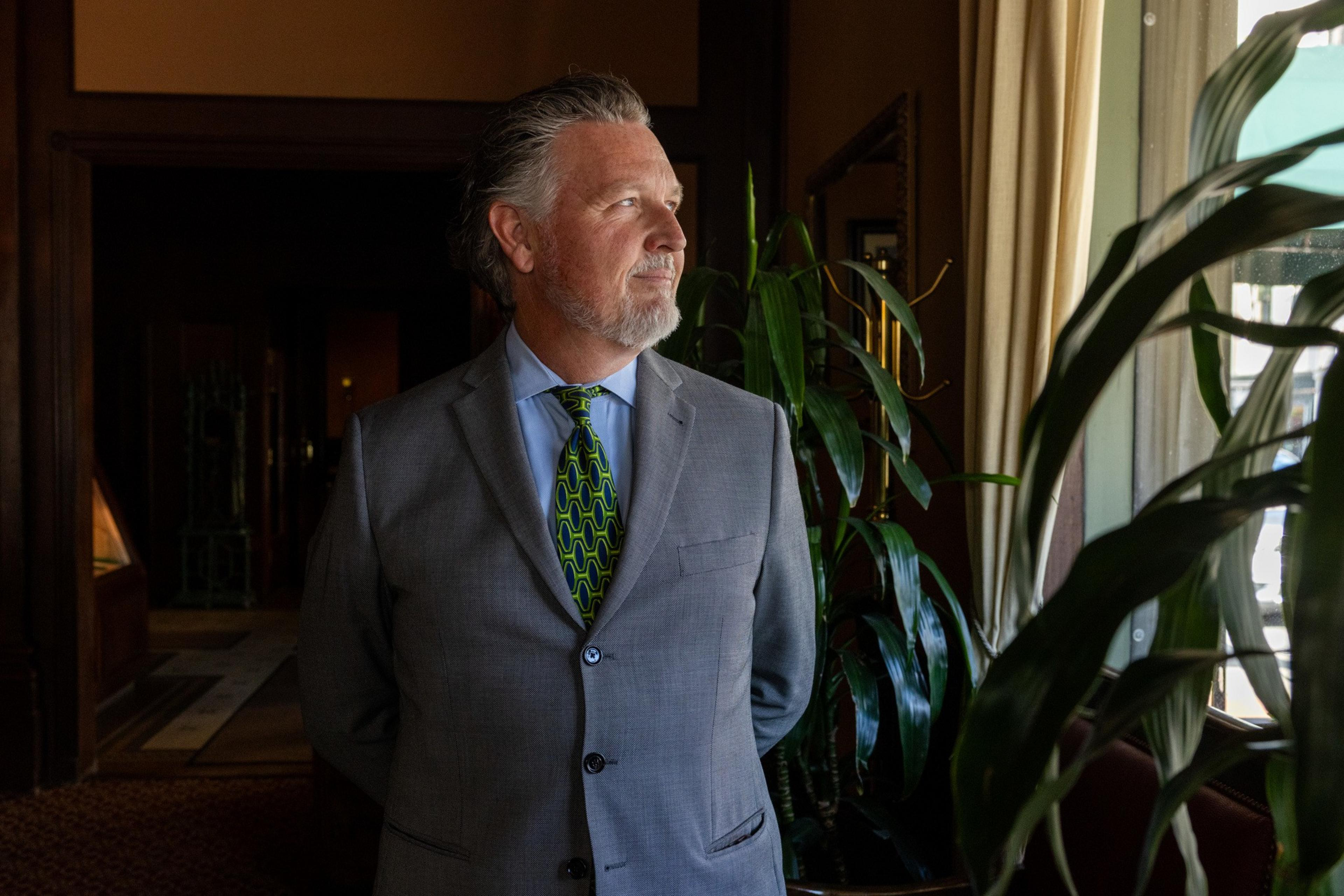 A man in a gray suit with a geometric tie stands indoors, looking out a window. Green plants are nearby, and the room has warm, dark tones.