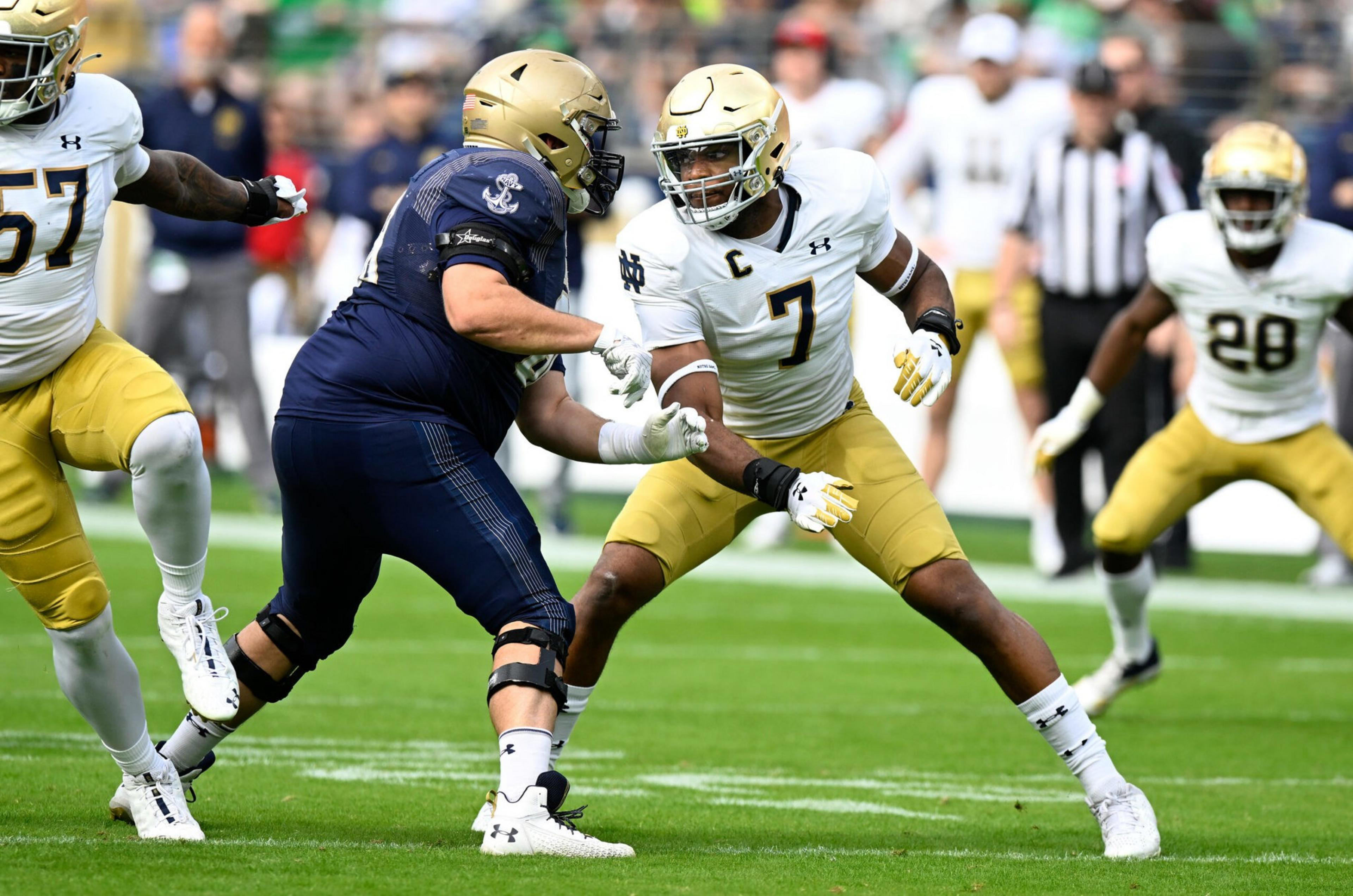 A football player in a white jersey with number 7 and gold helmet faces off against a player in a navy uniform during a game on green turf.