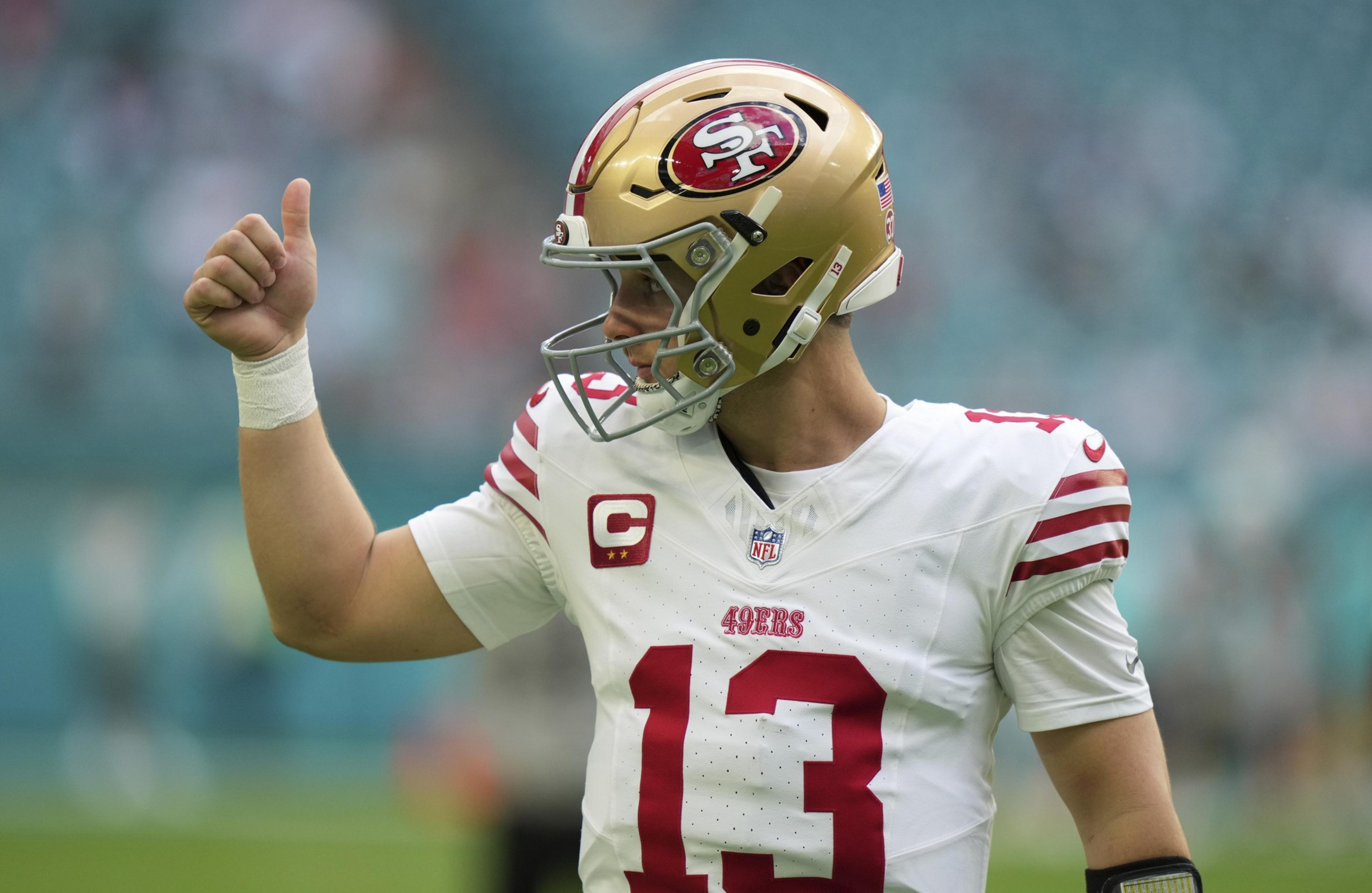 A football player wearing a gold helmet and white jersey with red details is giving a thumbs-up on the field. The background is a blurred stadium.