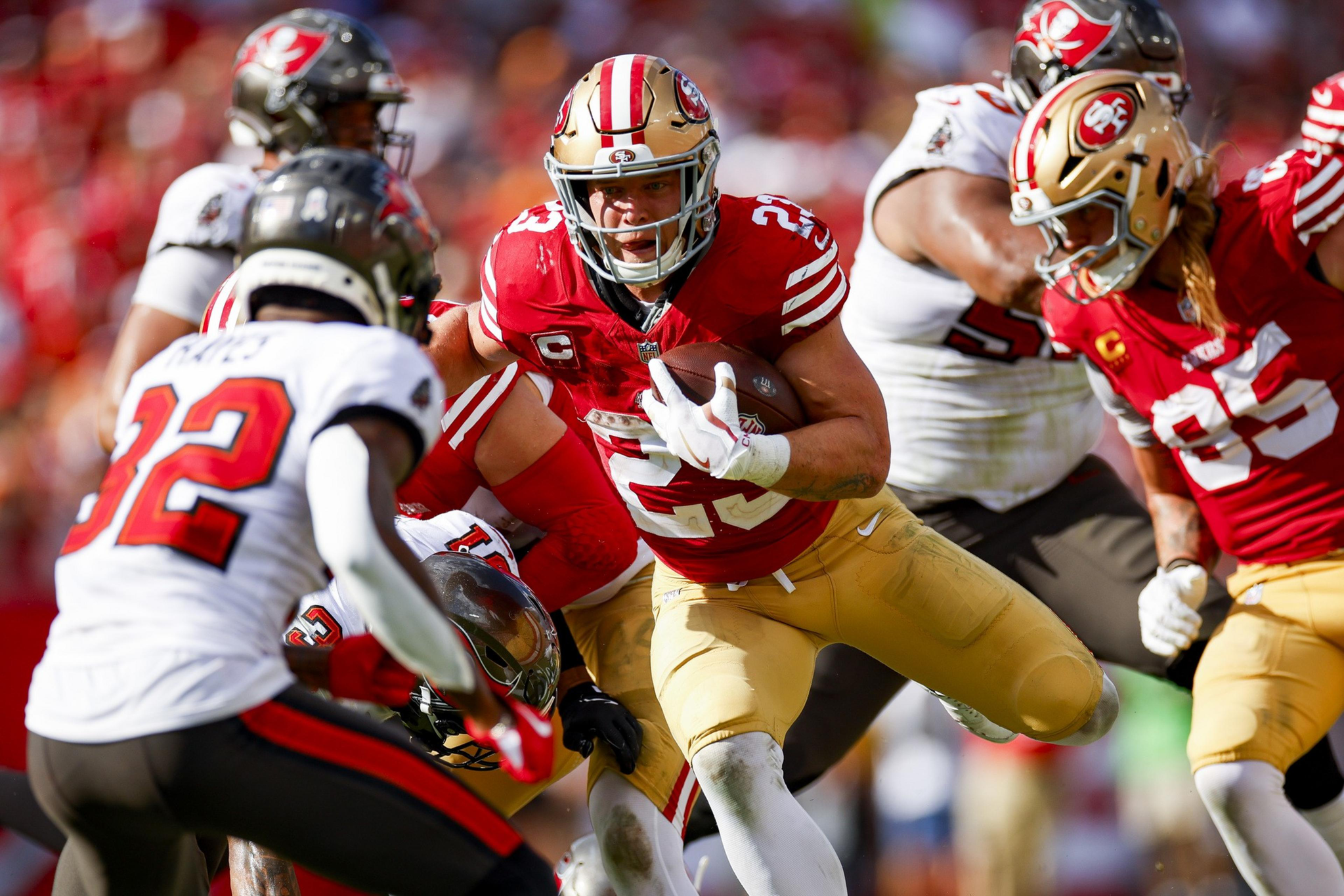 A football player in a red and gold uniform runs with the ball, dodging opposing players in white and black uniforms during a game.
