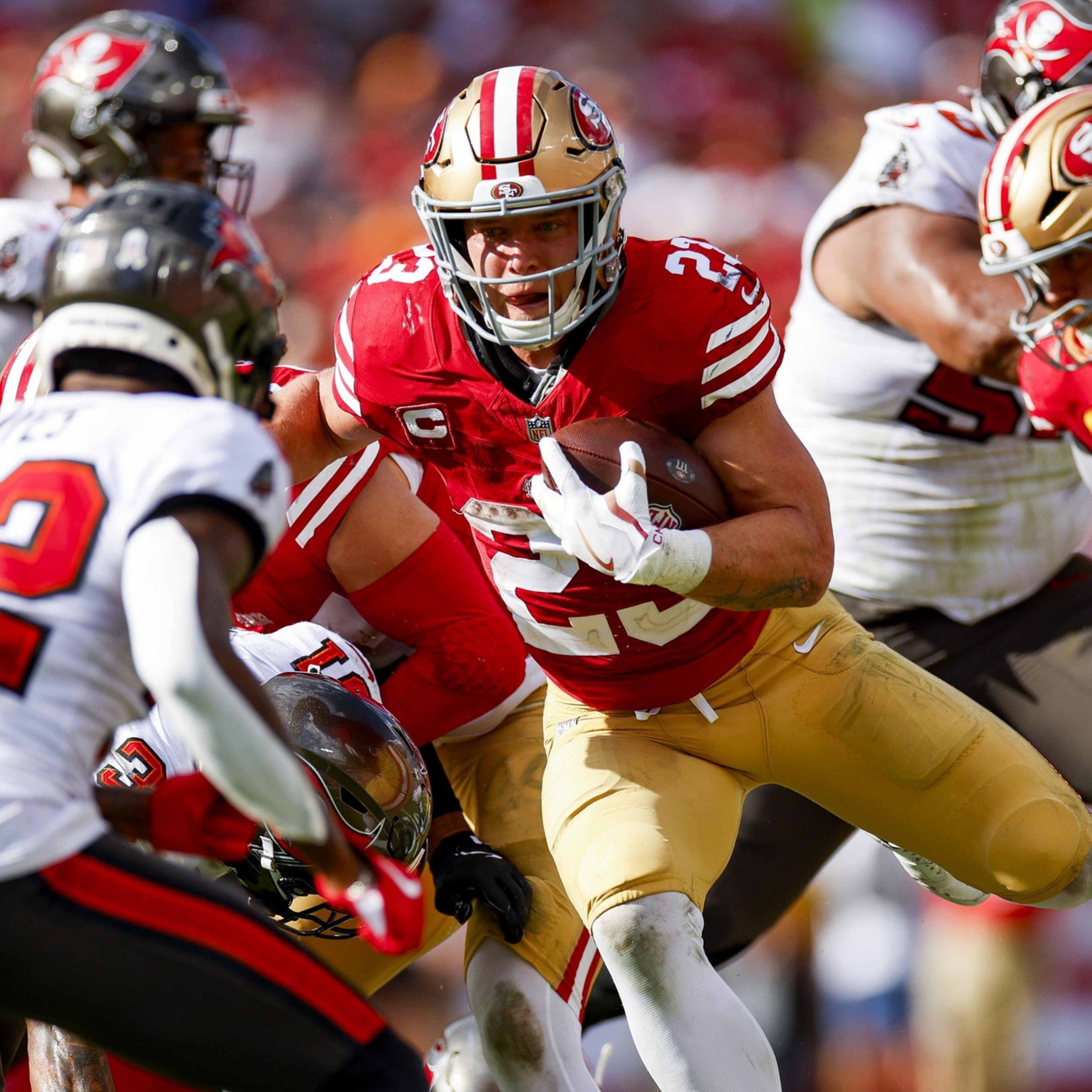A football player in a red and gold uniform runs with the ball, dodging opposing players in white and black uniforms during a game.