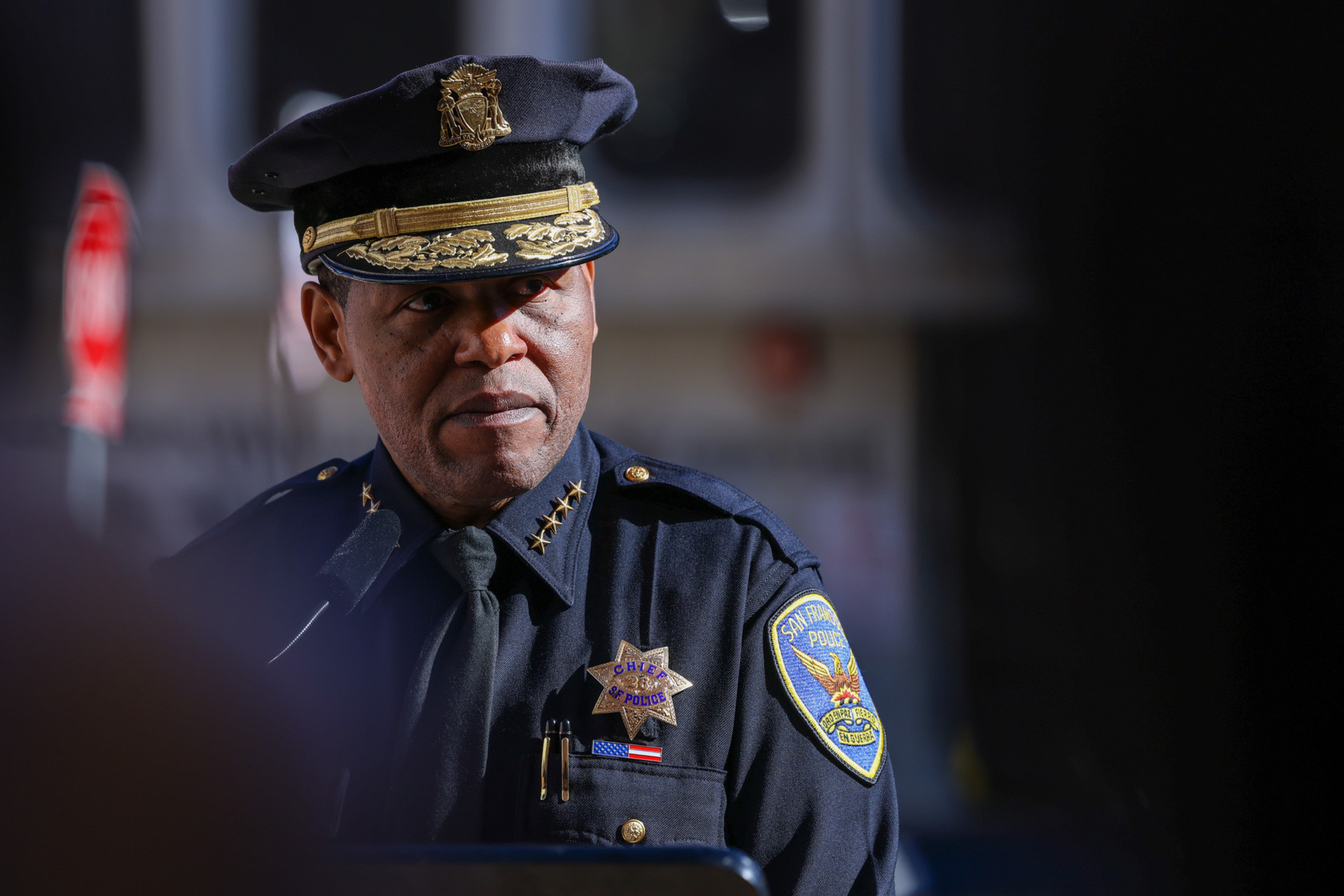 A police chief in uniform with a decorated hat and badges stands in partial shadow, looking intently to the side.