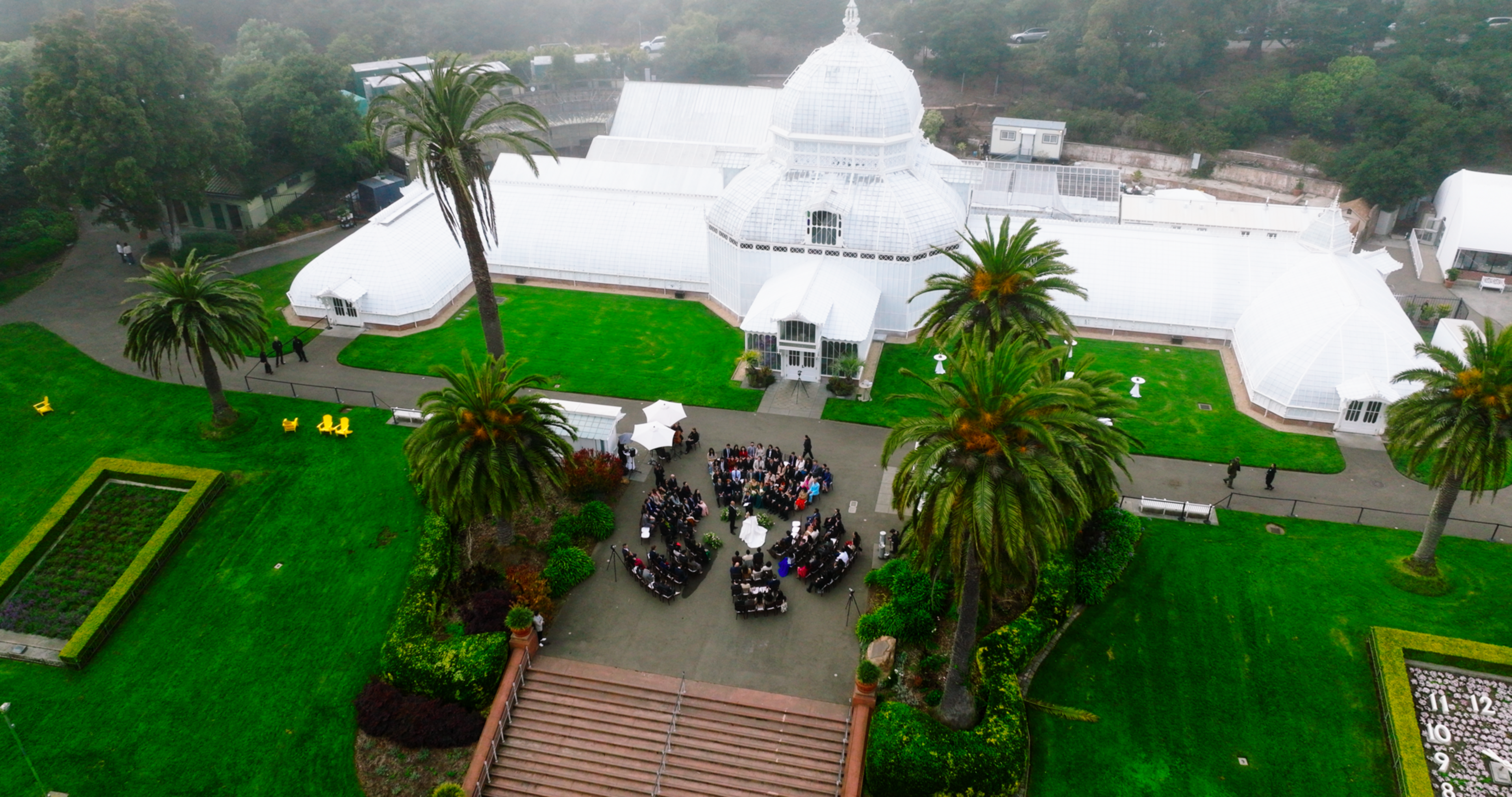 A group of people is gathered in a courtyard with palm trees and green lawns around a large white glass conservatory building.