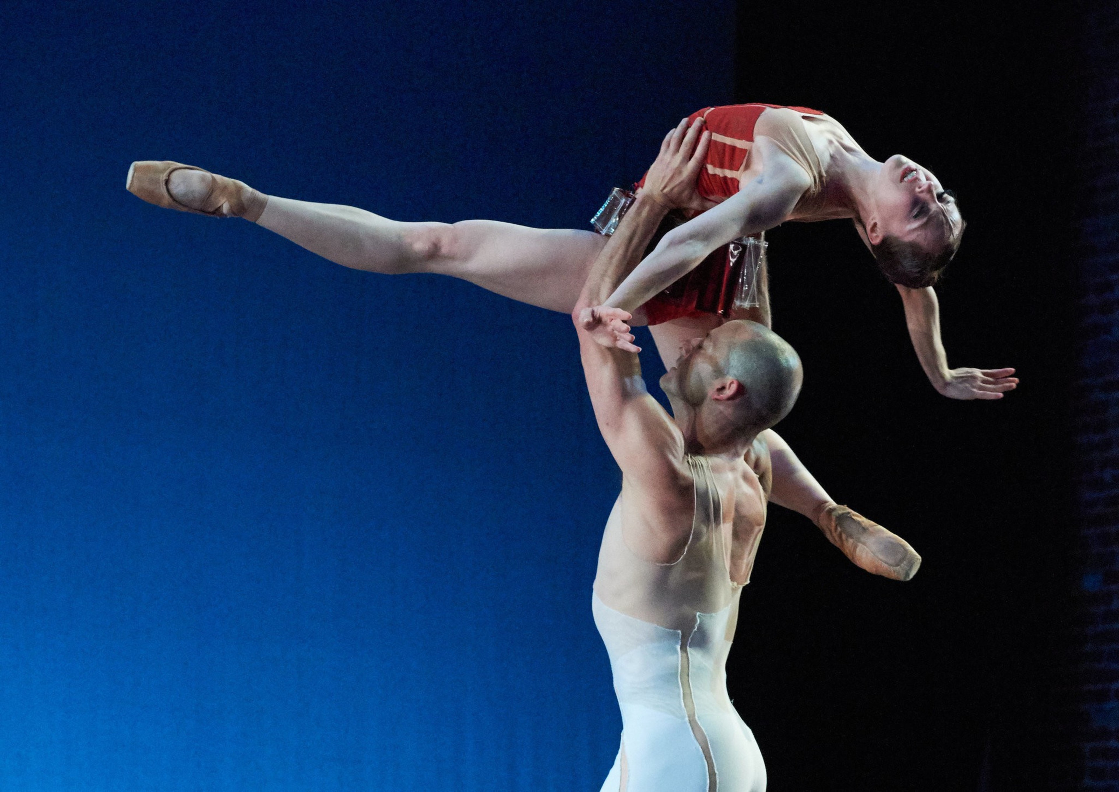 A male ballet dancer lifts a female dancer horizontally above his head as she stretches one leg straight out and arches her back.