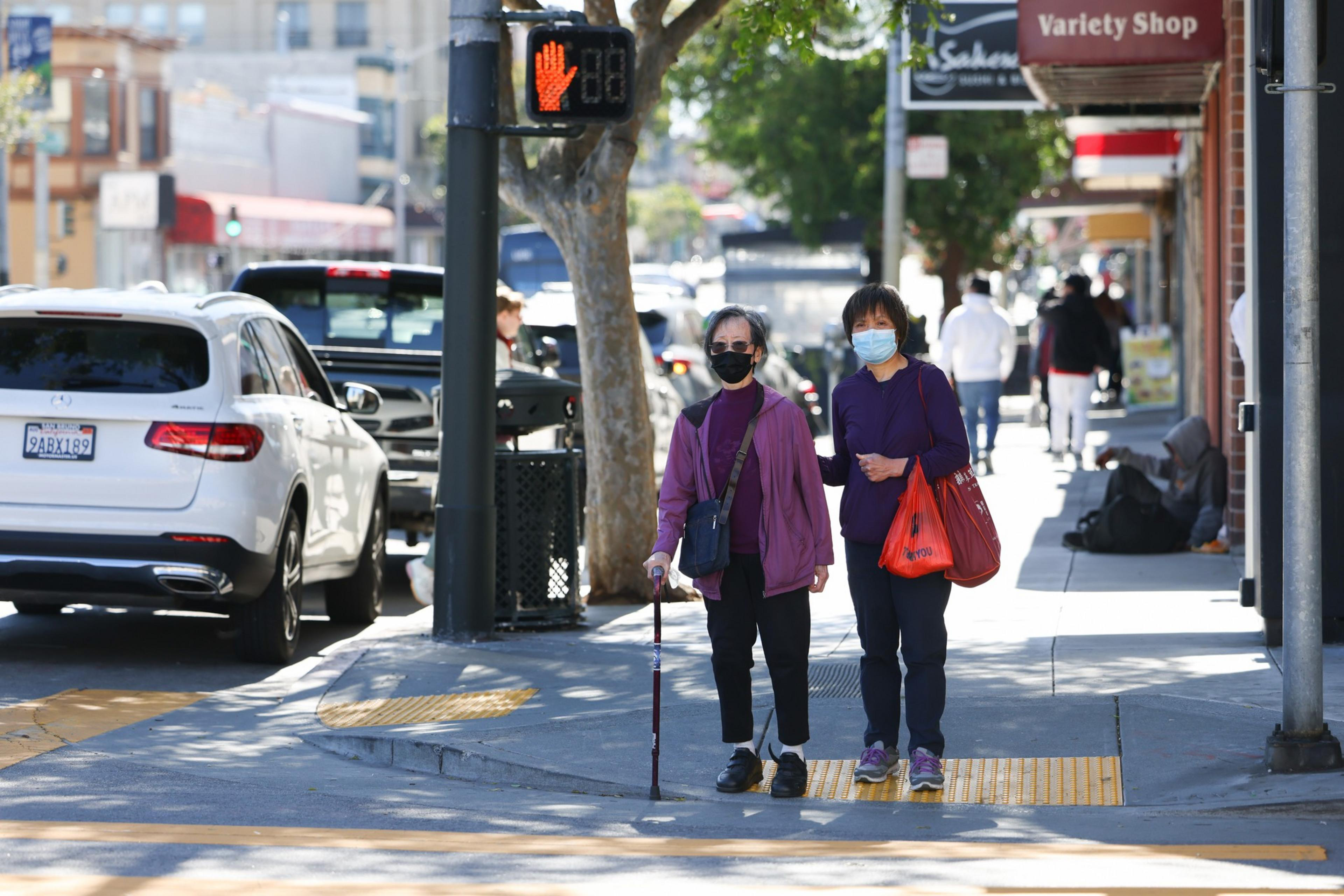 Two women wearing masks cross a street together, one using a cane, under a traffic signal displaying a red hand to stop.