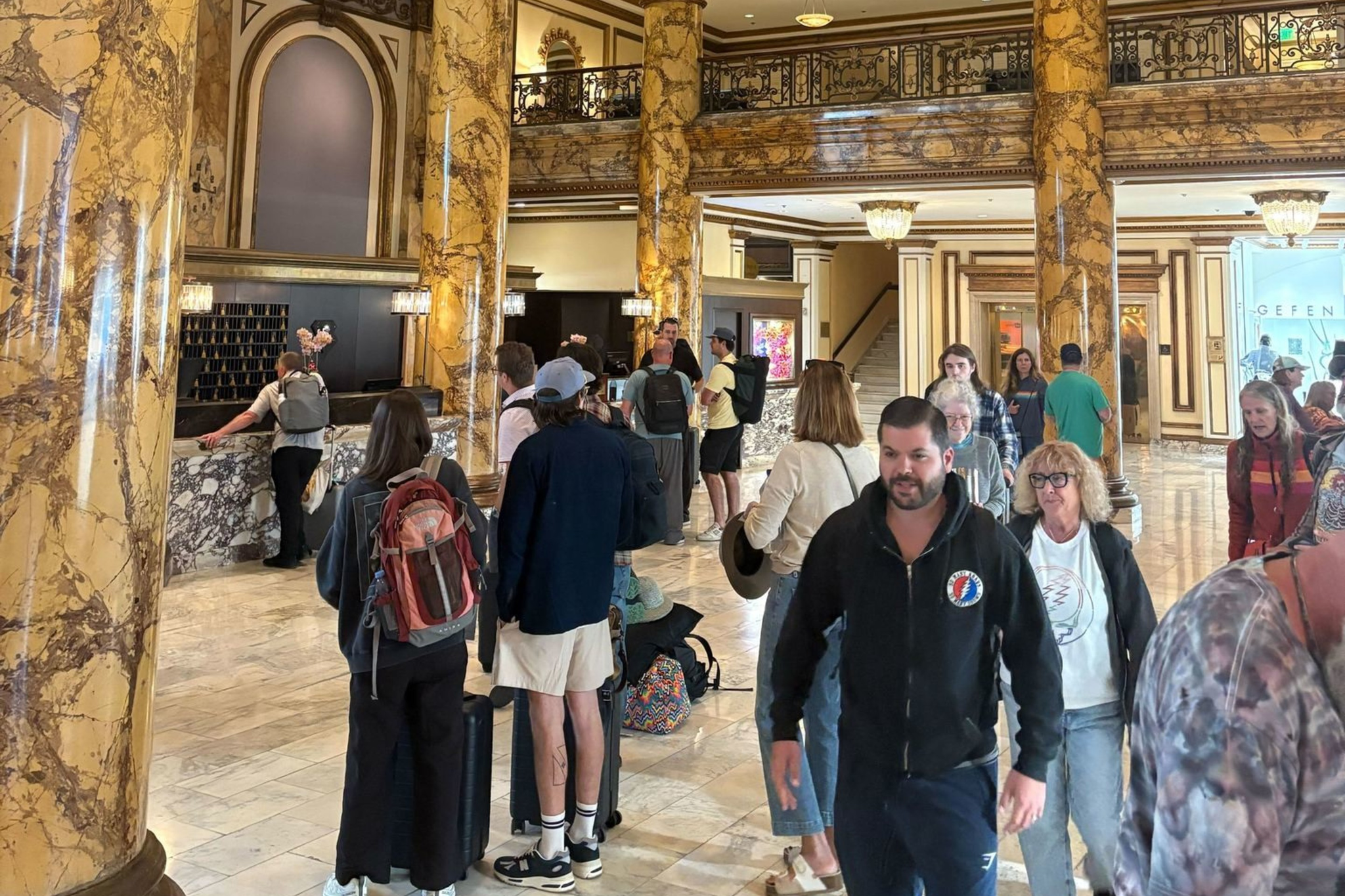 A grand lobby with tall marble columns, ornate ceiling, and chandelier, filled with people carrying luggage and standing in line.