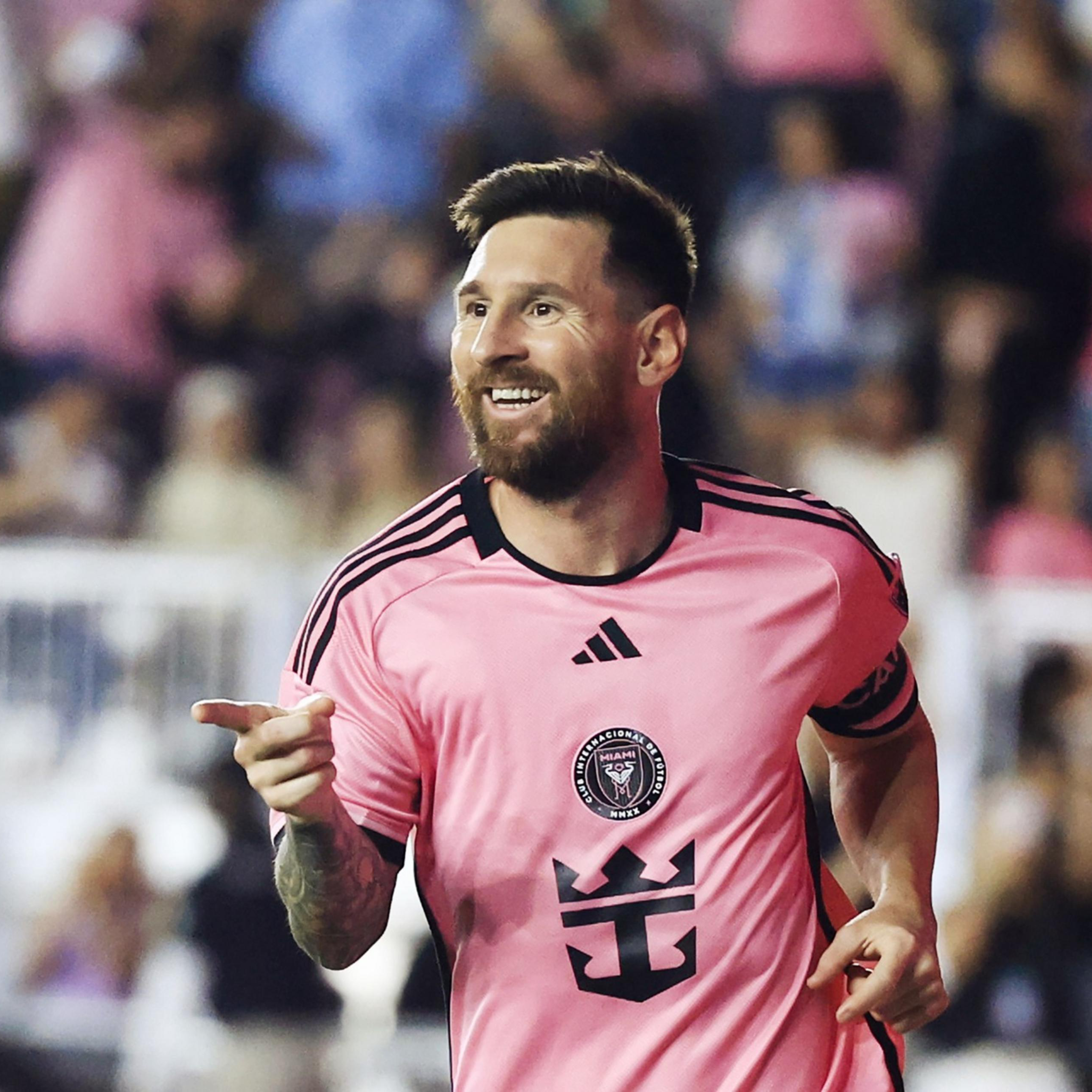A soccer player in a pink jersey is smiling and pointing, with an enthusiastic crowd in the background.