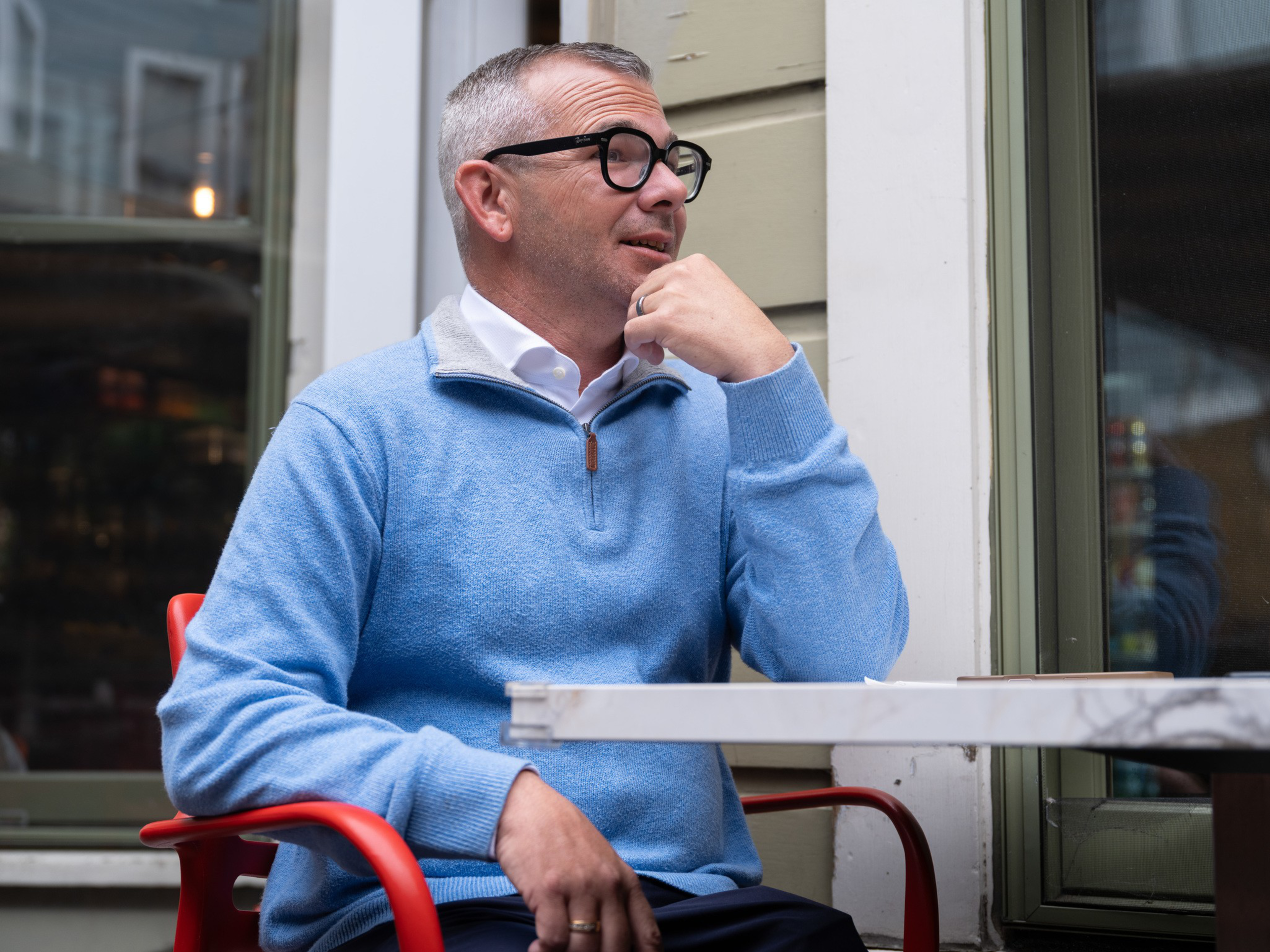 A man with short gray hair and glasses sits at an outdoor café table, wearing a blue sweater over a white collared shirt.