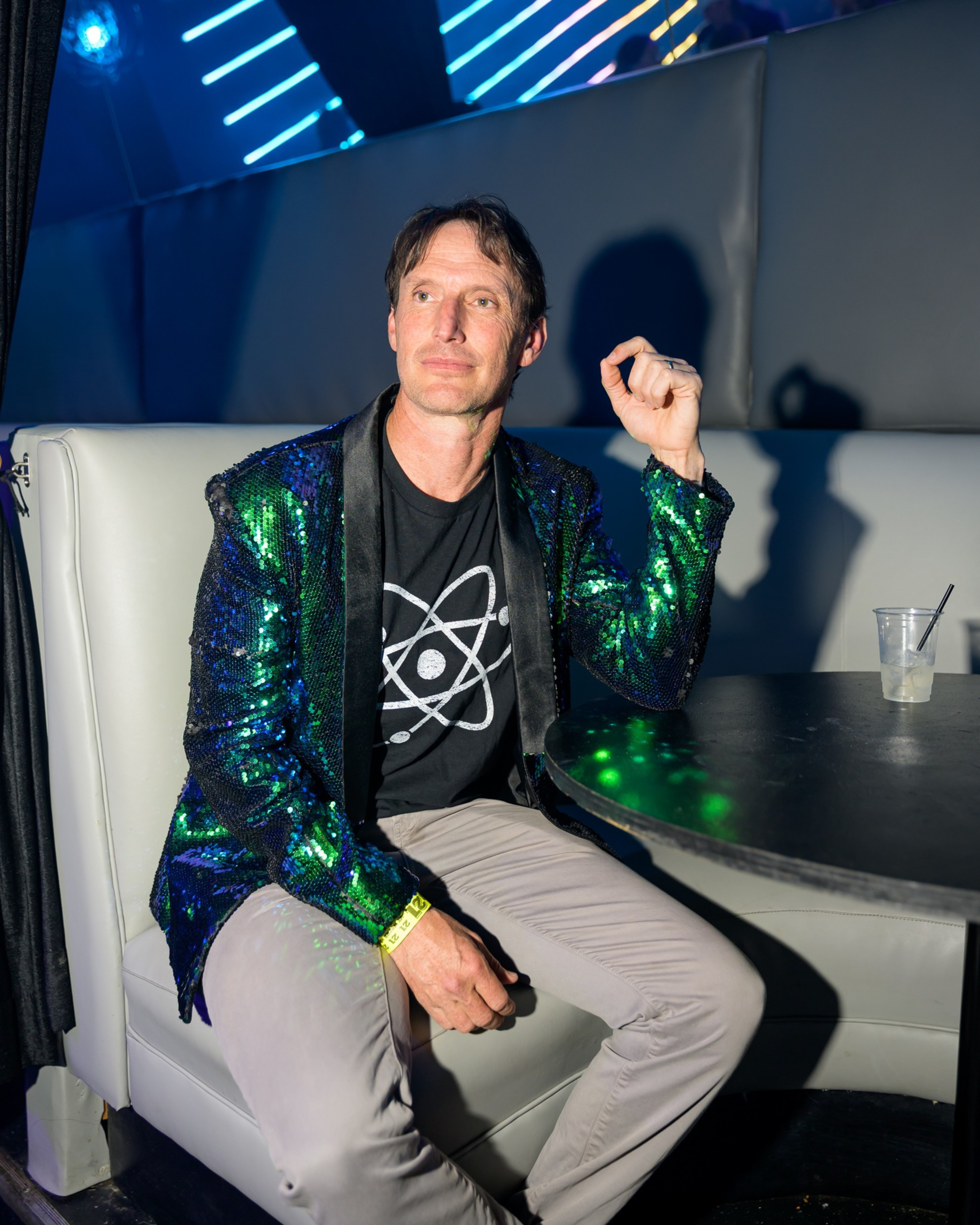 A person in a green sequin jacket and atomic logo shirt sits in a dimly lit booth with a drink on the table. Neon lights illuminate the background.