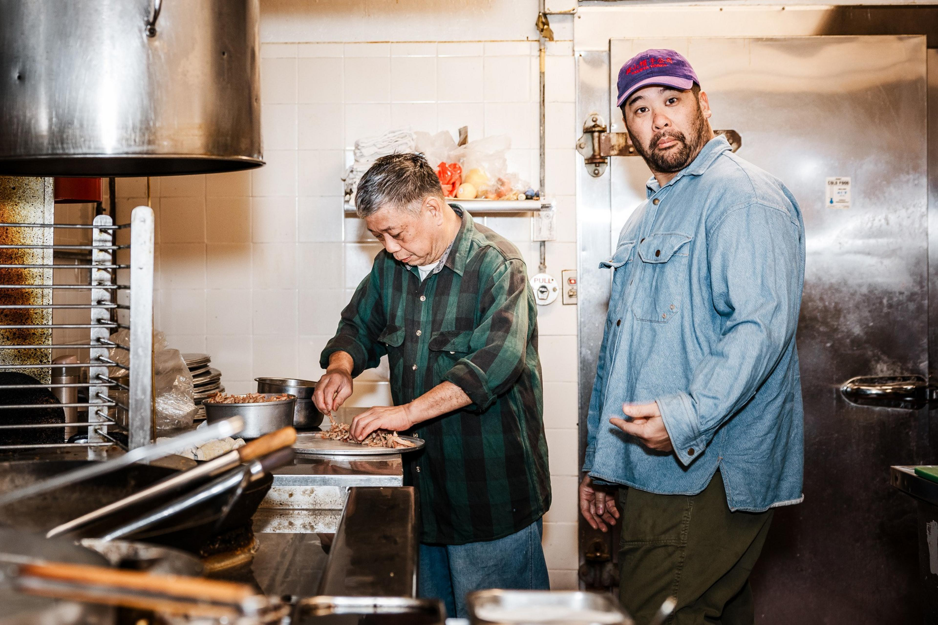 Two men are in a kitchen; one is chopping food on a plate, while the other looks at the camera with a surprised expression. They're surrounded by cooking tools.