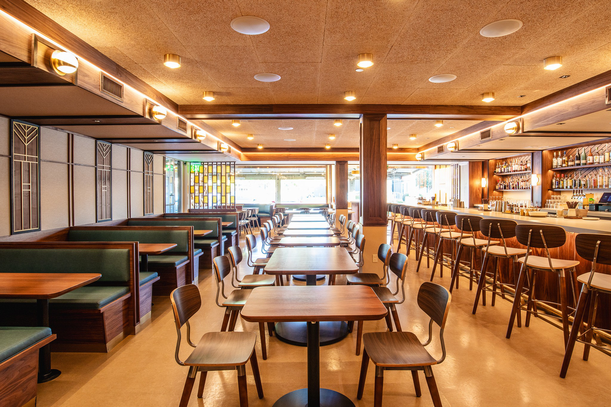 A brightly lit restaurant interior with wooden tables, green cushioned booth seating on the left, and high bar stools along a stocked bar on the right.