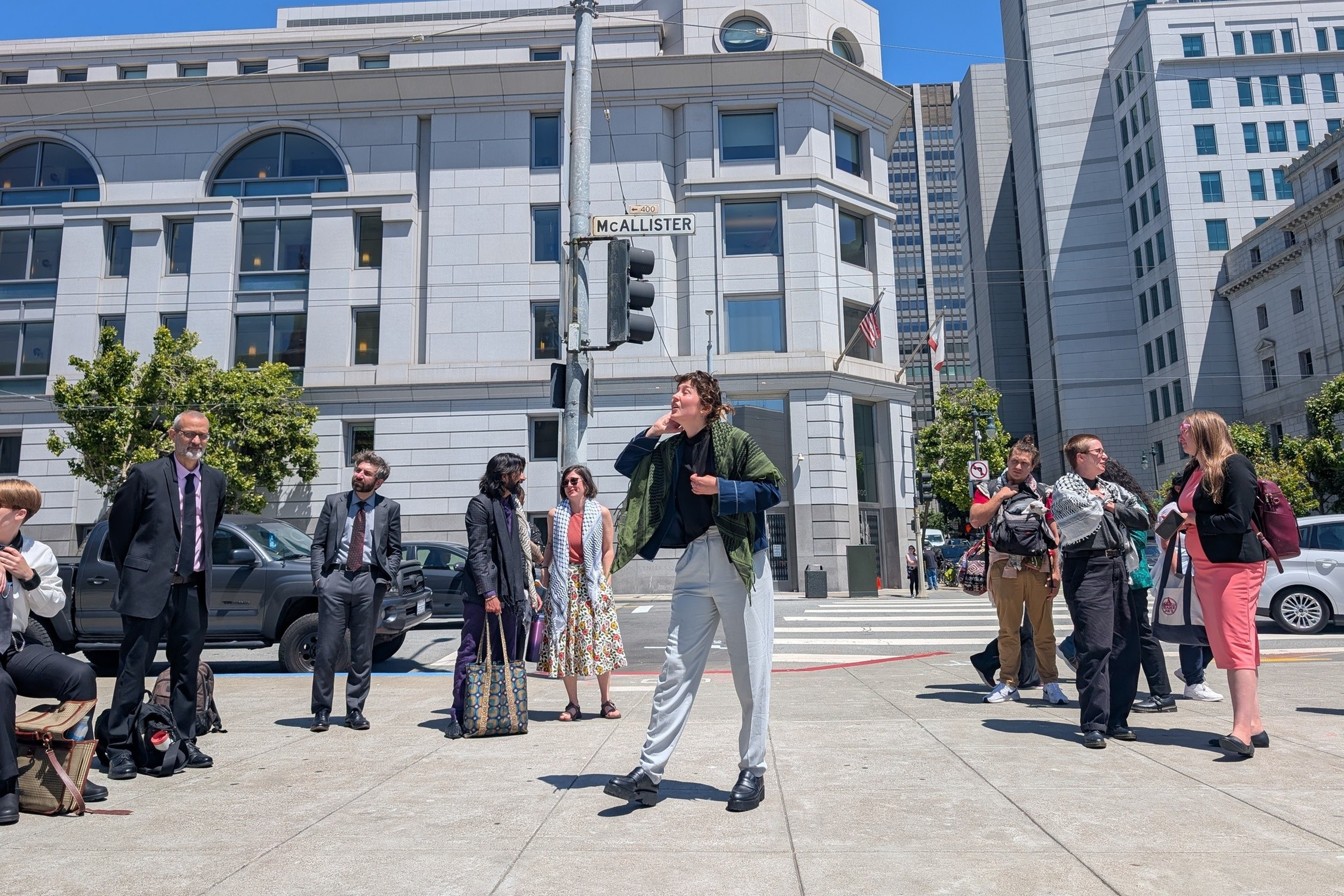 A group of people stand and wait at a street corner under a blue sky near a "McALLISTER" sign with tall buildings in the background.