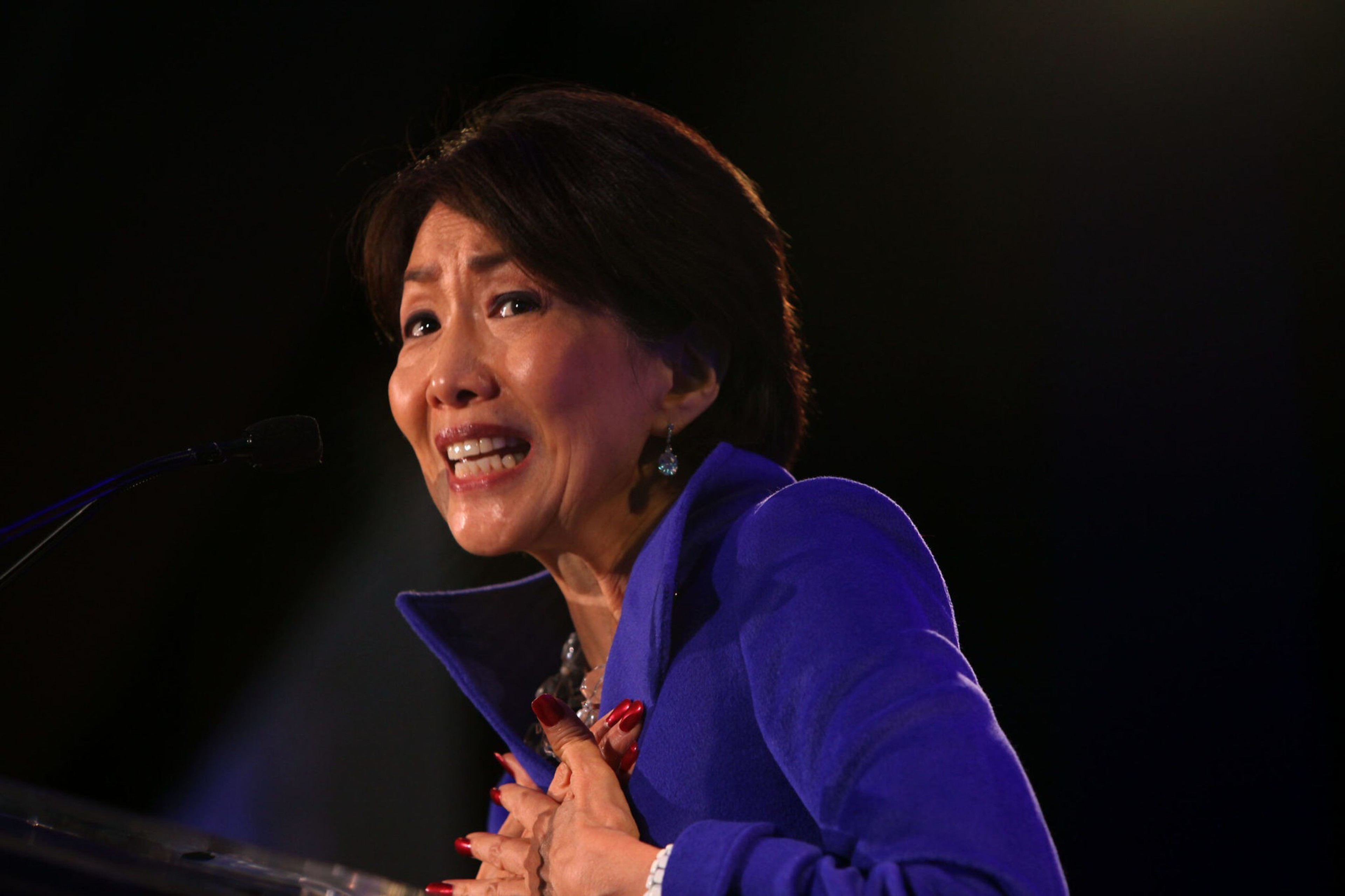 A woman in a vibrant blue jacket speaks passionately at a microphone, her hands clasped near her chest and an expressive look on her face.