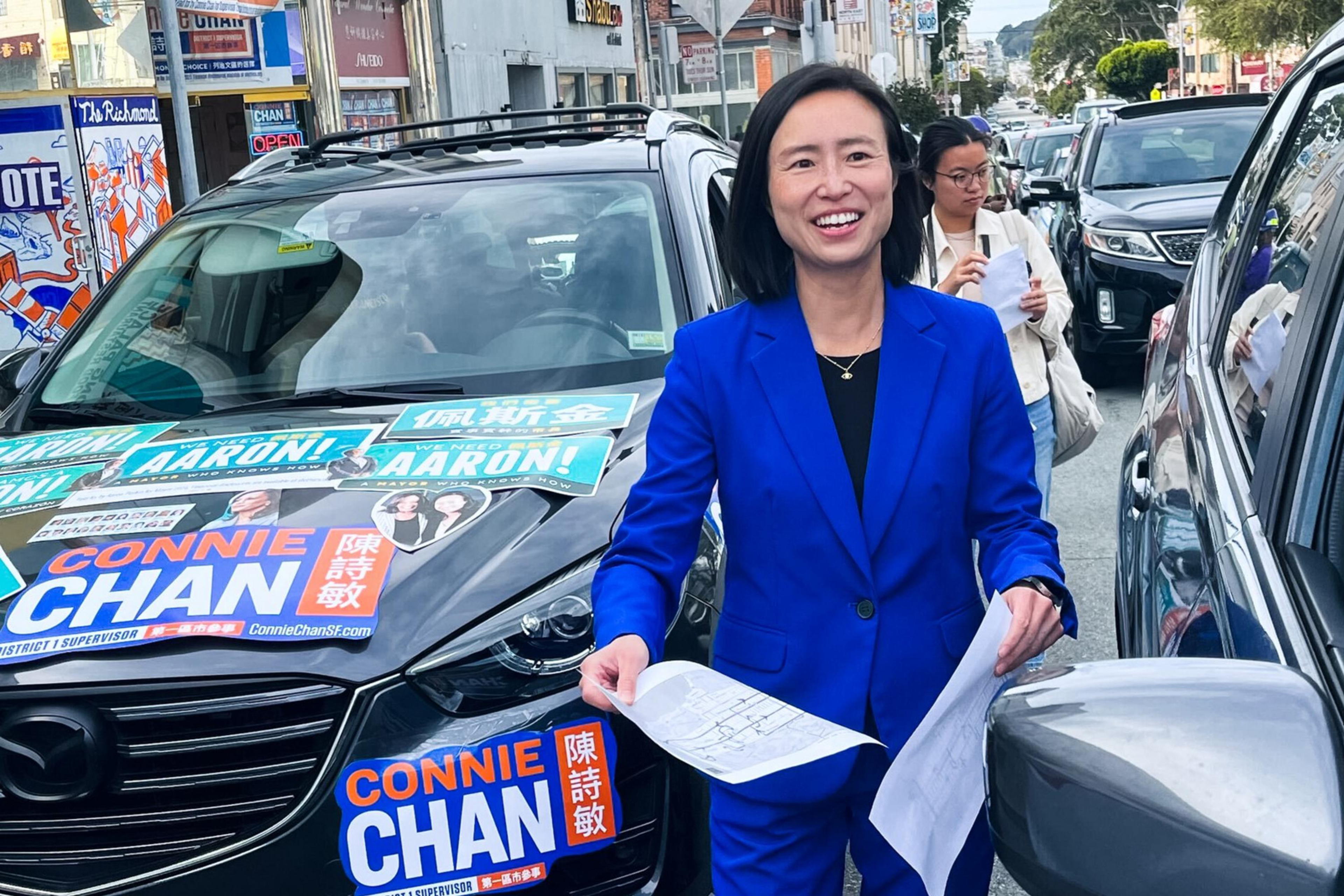 A person in a blue suit stands between two parked cars on a street. The car on the left is covered with campaign posters and signs. The person smiles while holding papers.