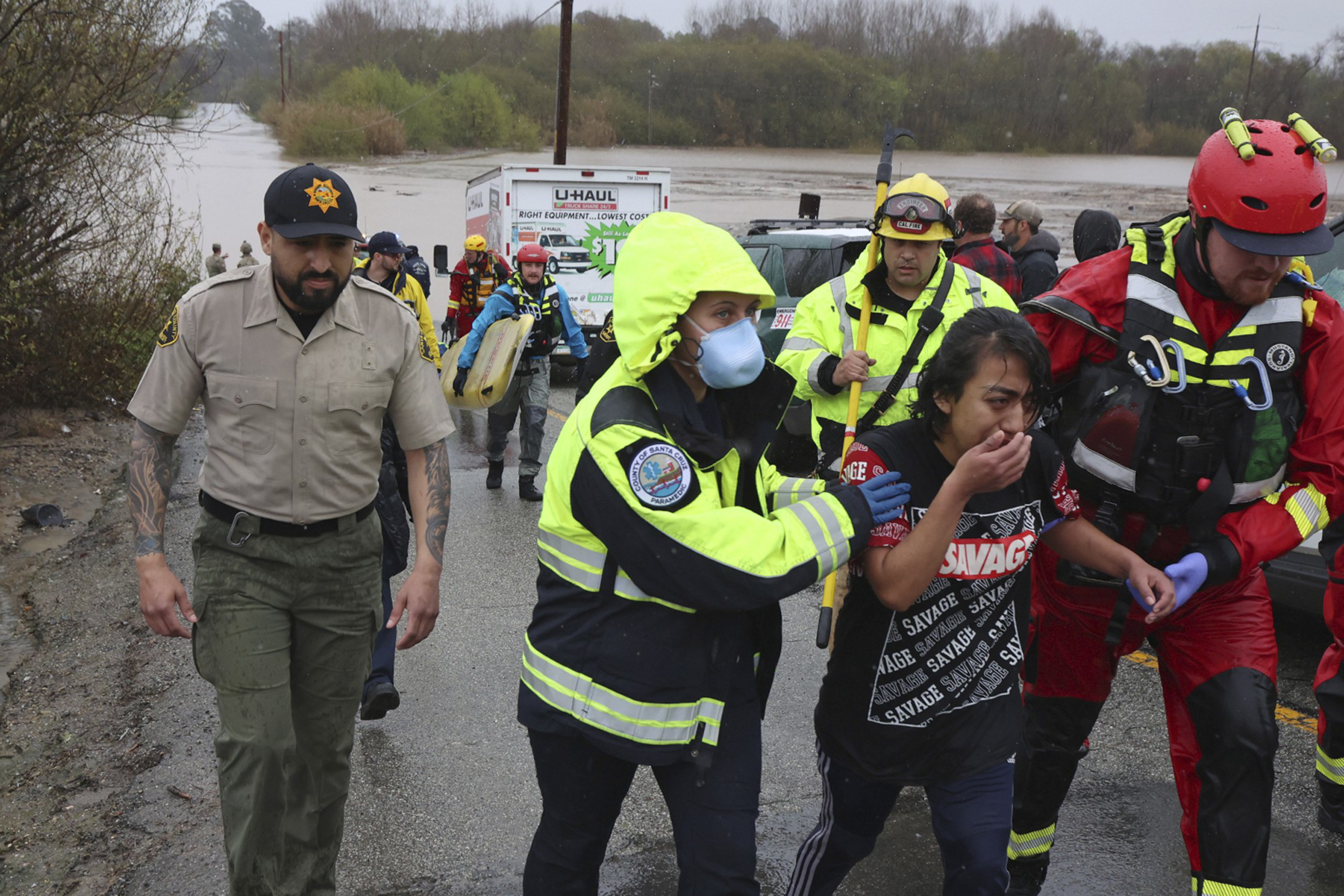Rescue workers in rain gear assist a distressed woman near a flooded area, with emergency vehicles and other responders in the background.