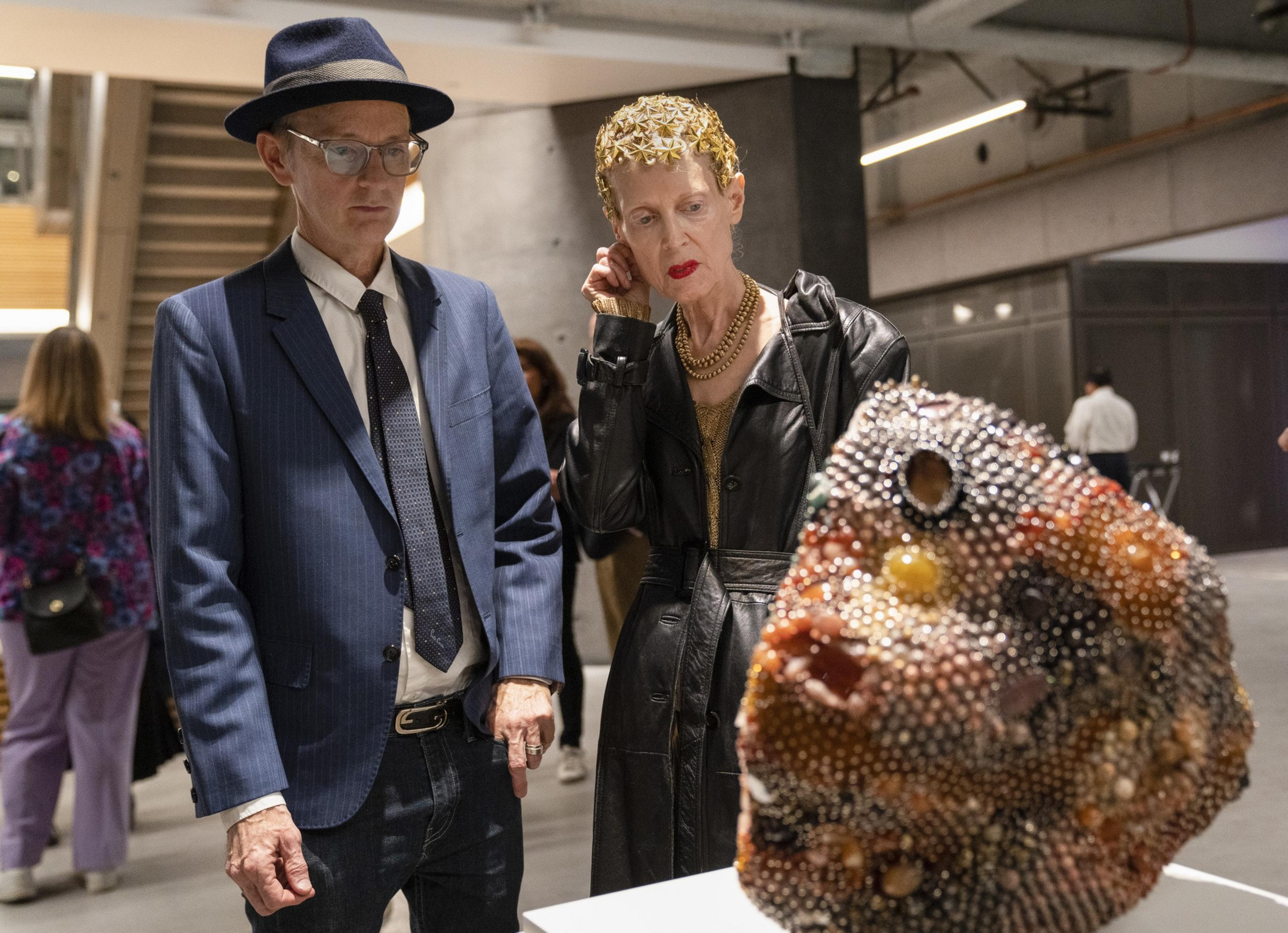 A man in a blue suit and hat stands next to a woman in a black coat, gazing at a textured, colorful sculpture. The background shows an indoor art gallery space.