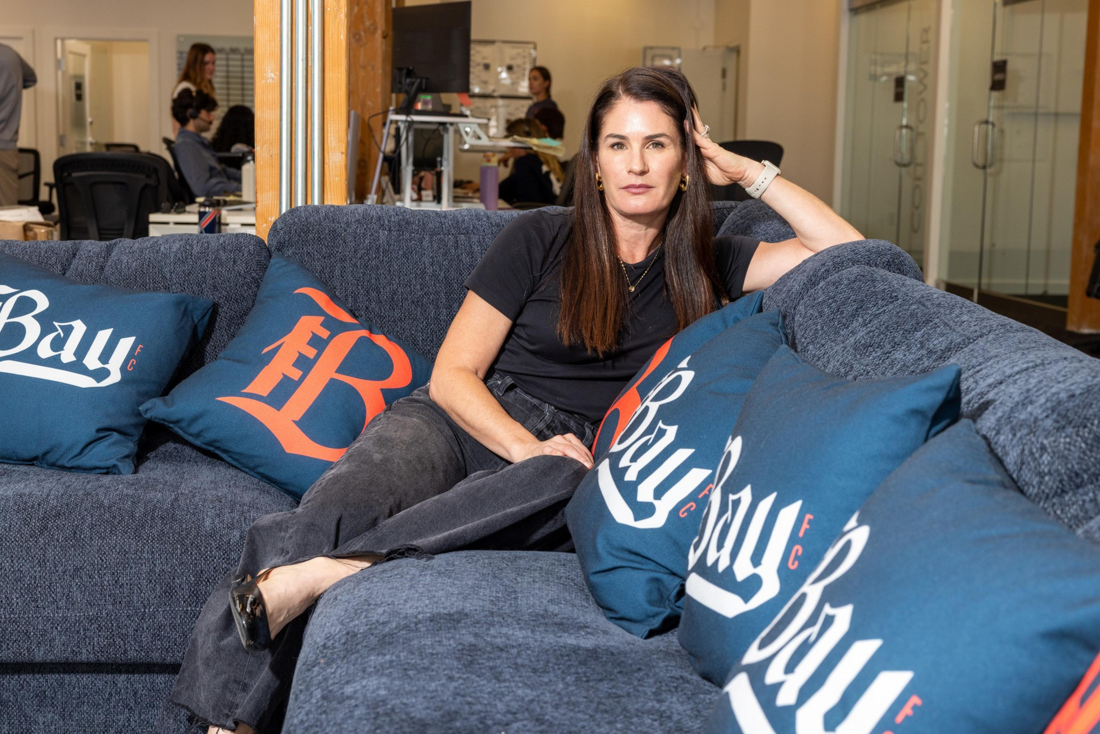 A woman with long brown hair wearing a black shirt and pants lounges on a blue couch with blue pillows labeled "Bay FC."