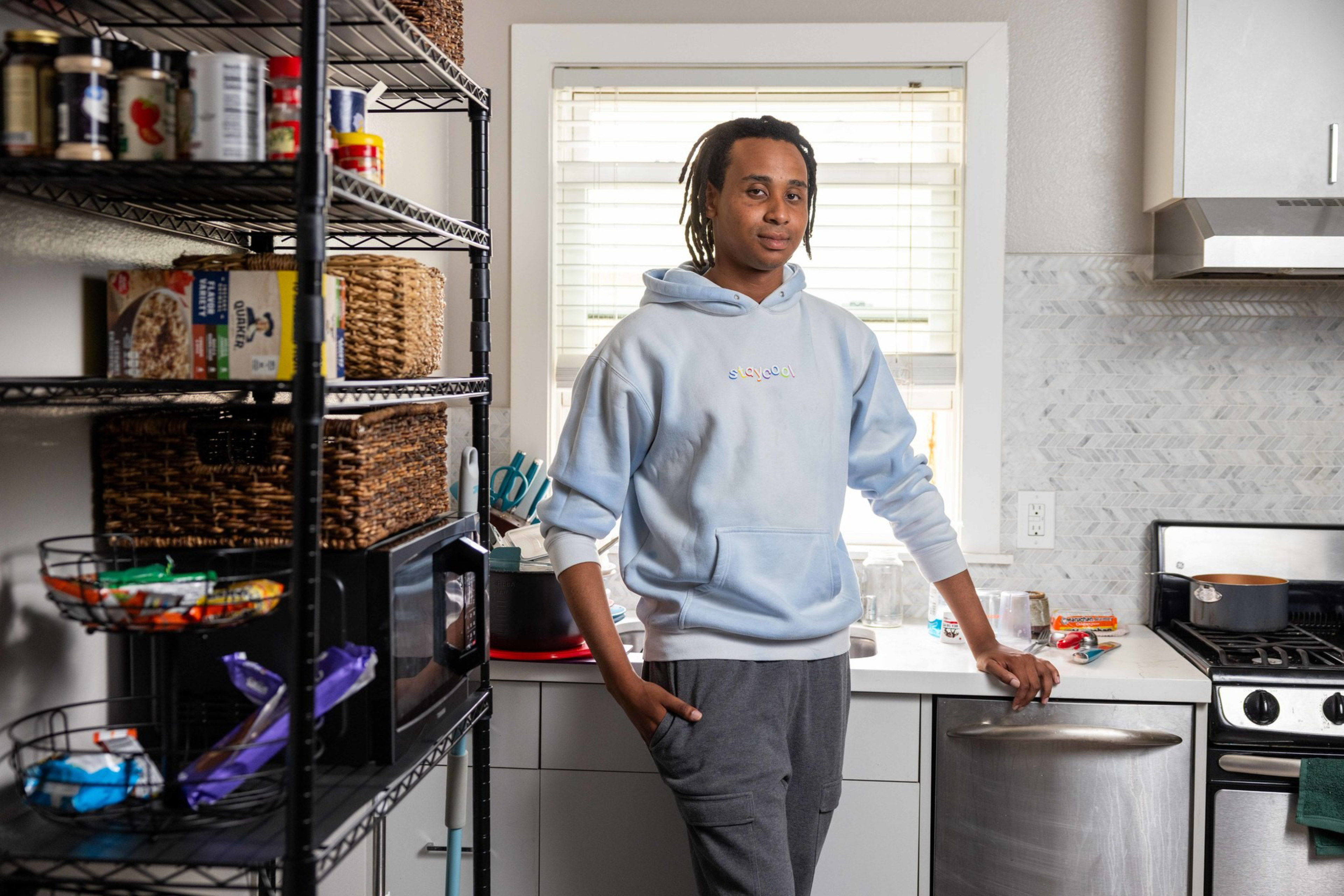 A young person in a light blue hoodie stands in a kitchen near a window, with a metal shelving unit full of food and a stove with a pot.