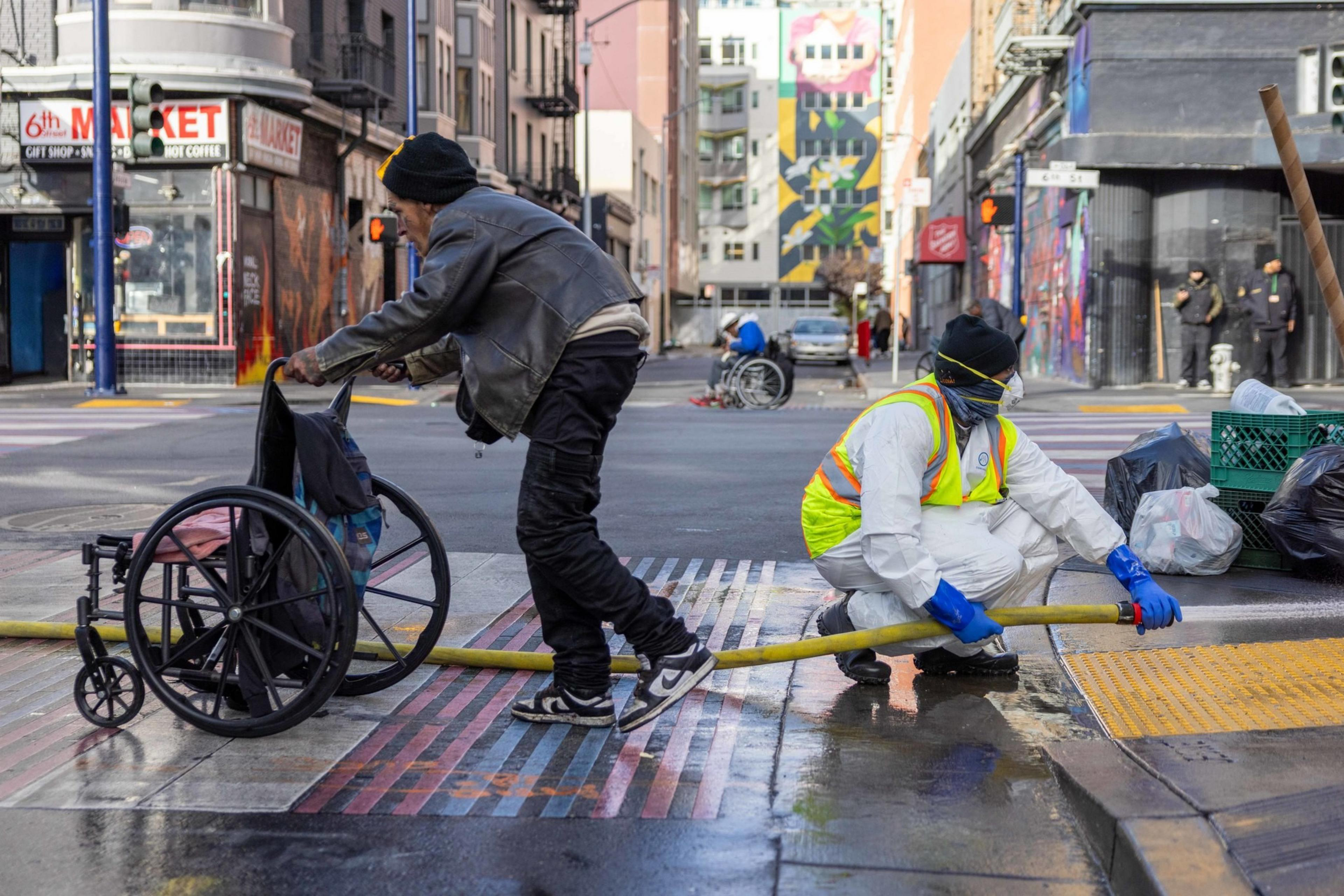 A man pushes a wheelchair across a wet street while another person in protective gear and a high-visibility vest cleans the sidewalk with a hose.