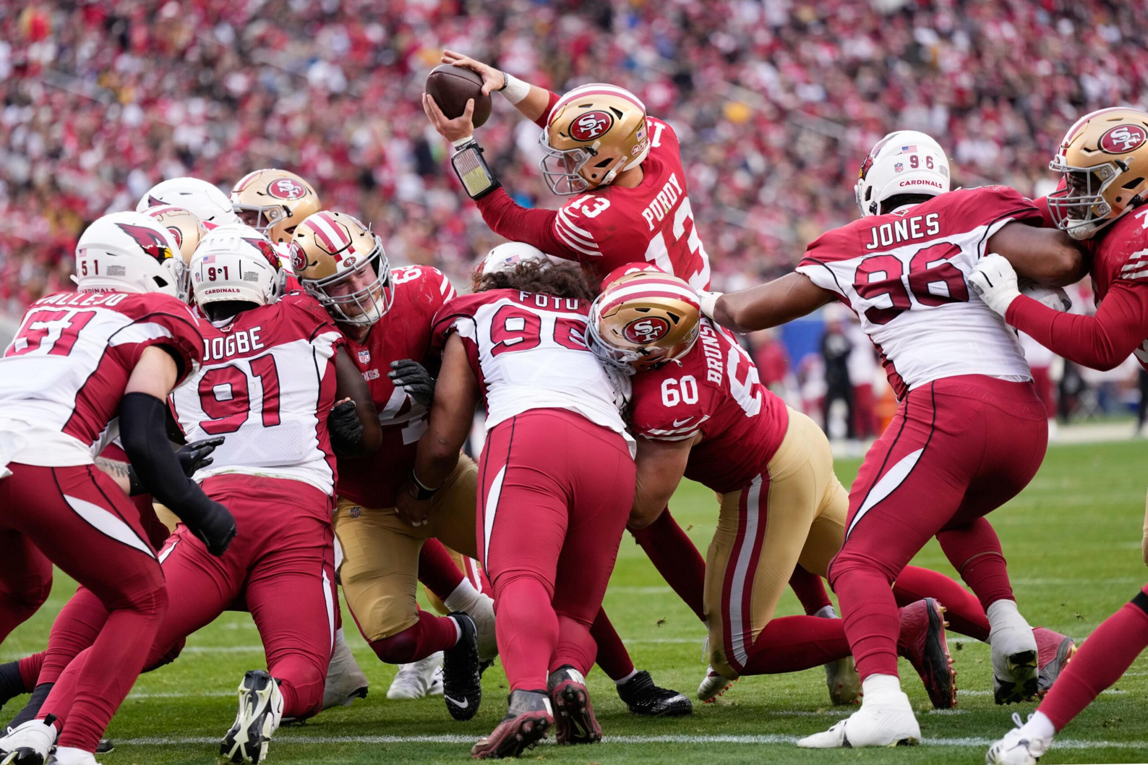A football quarterback in a red and gold uniform attempts to throw while being tackled by multiple players in white and red jerseys on a green field.