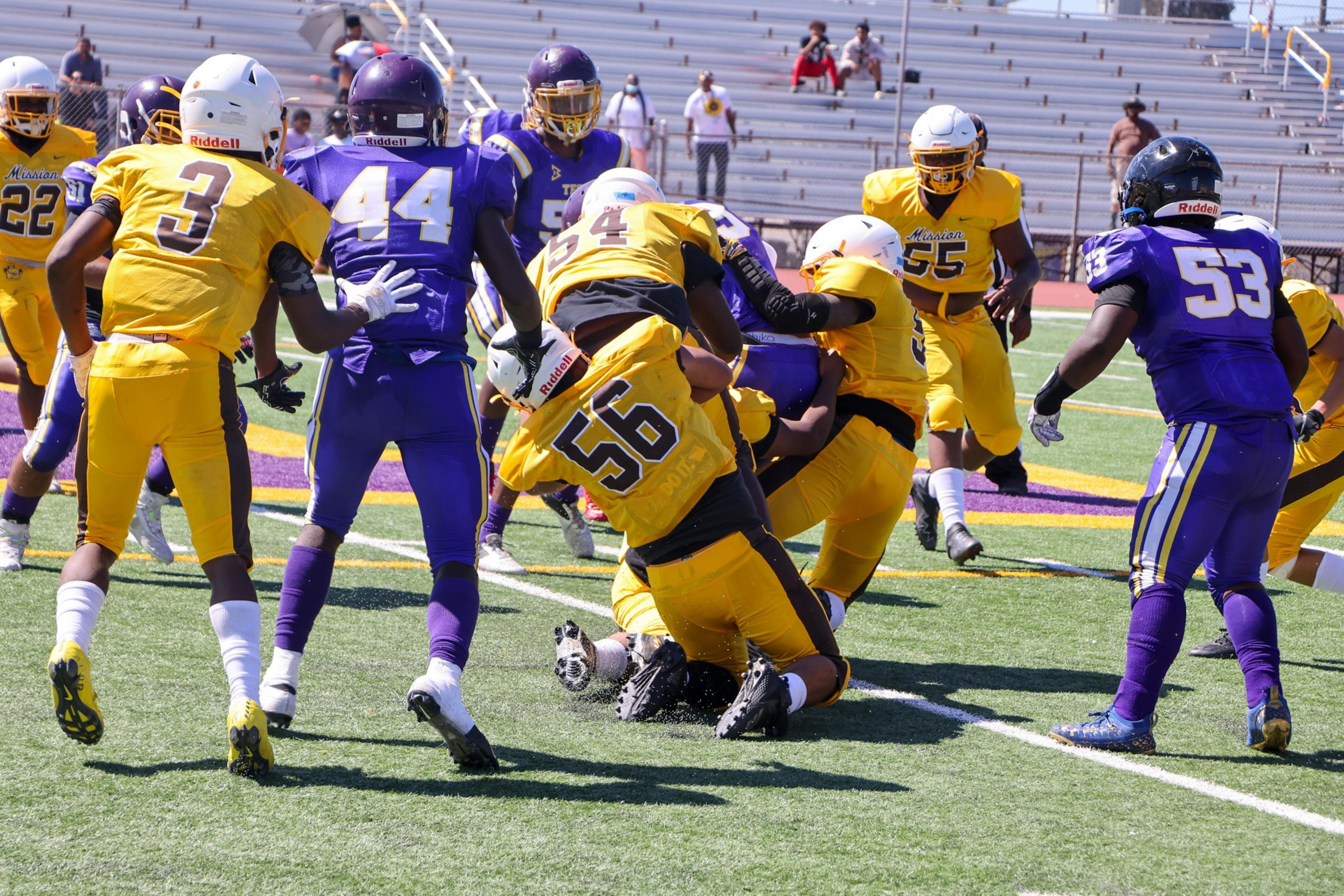 Football players in yellow and purple uniforms clash on a green field as one yellow player tackles a purple player holding the ball near the end zone.