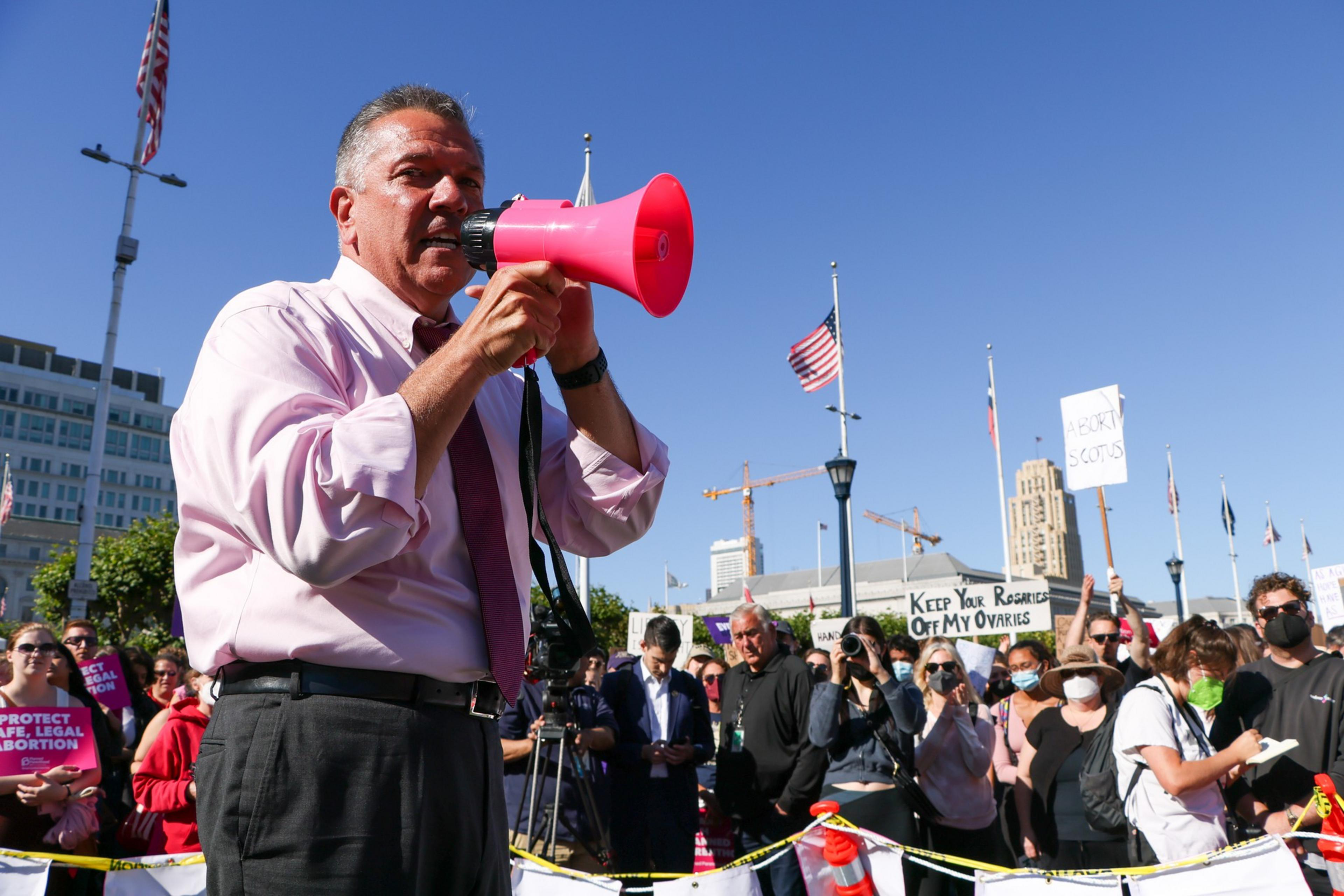 A man in a pink shirt speaks into a red megaphone at a protest, with people holding signs about abortion rights in the background.