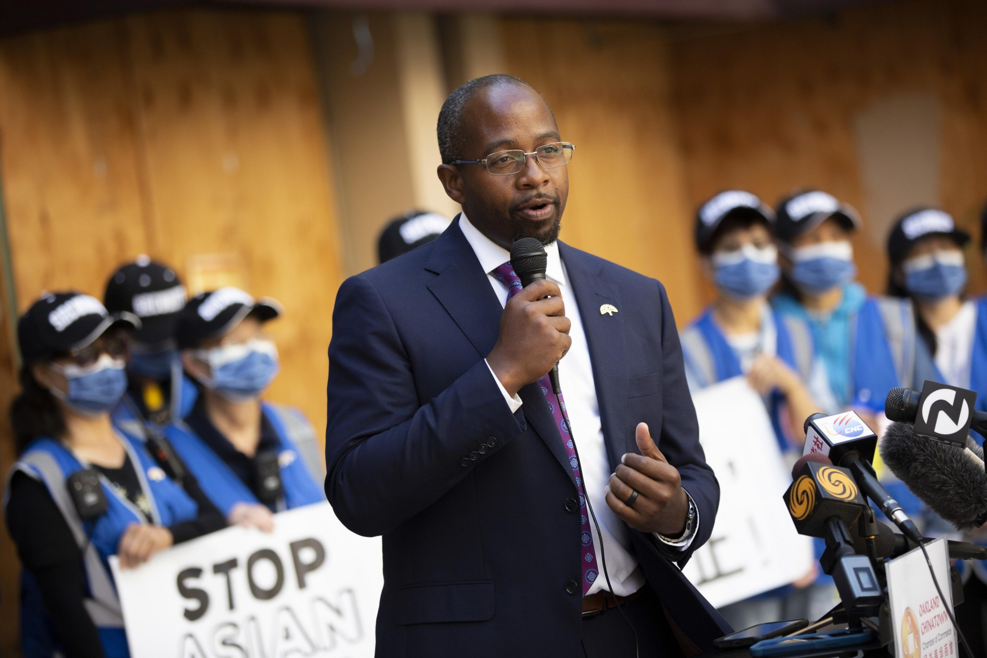 A man in a suit speaks into a microphone at a podium with microphones bearing news logos. Behind him, masked individuals hold signs reading "STOP ASIAN HATE."
