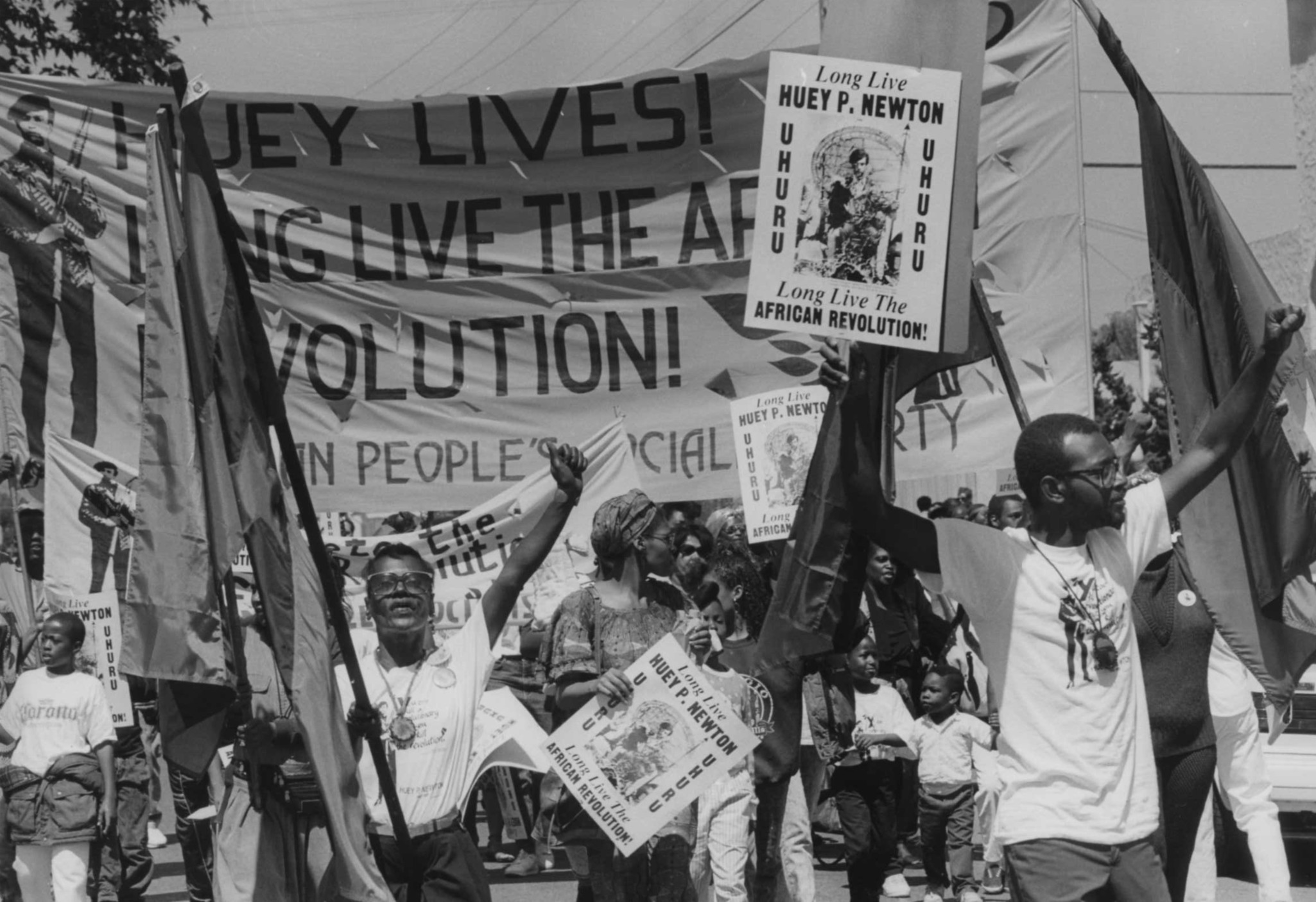 People march holding signs and flags celebrating Huey P. Newton and the African revolution, with a large banner reading "Huey lives! Long live the revolution!"