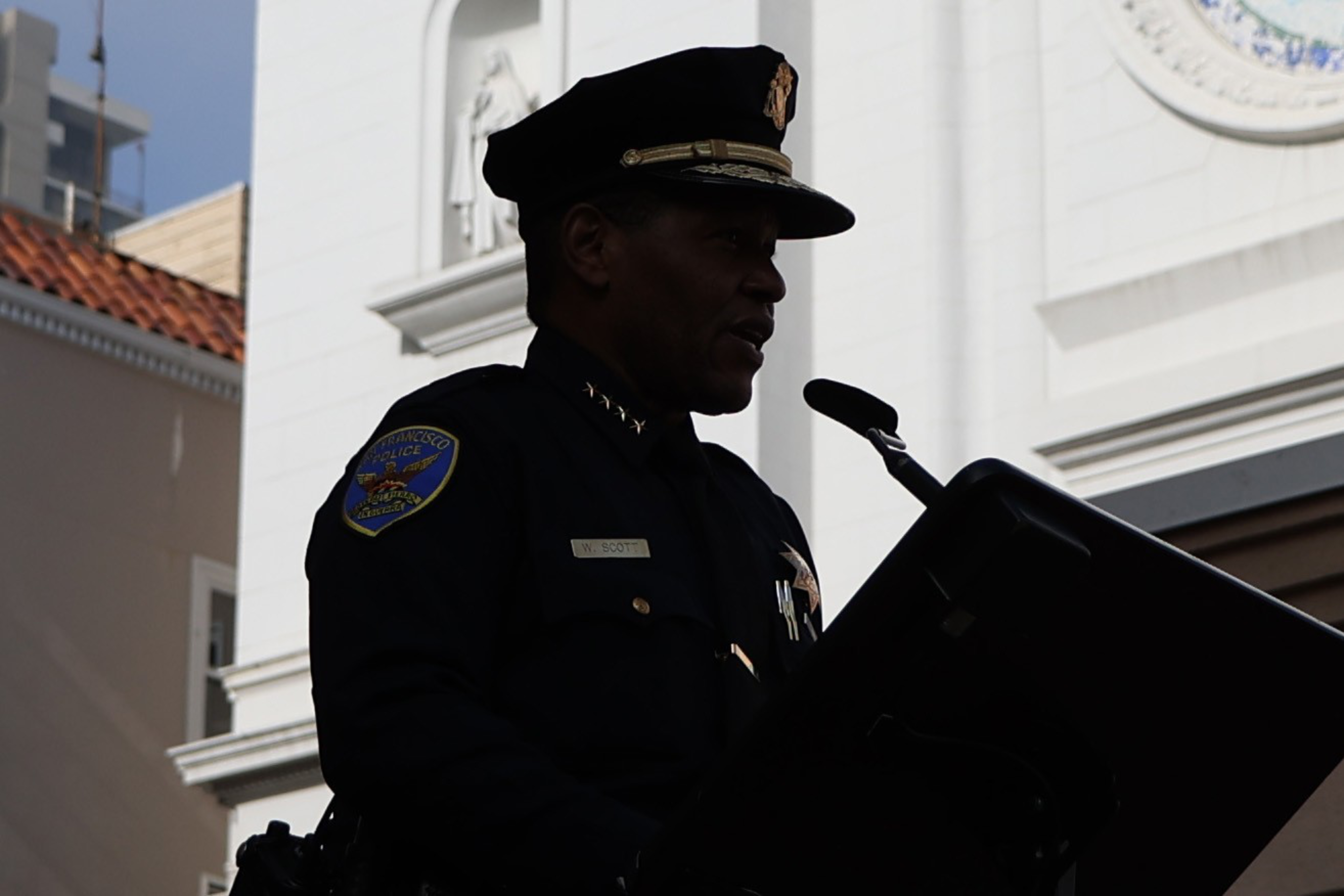 A police officer in uniform is speaking at a podium with a microphone, with a building featuring a statue and decorative elements in the background.