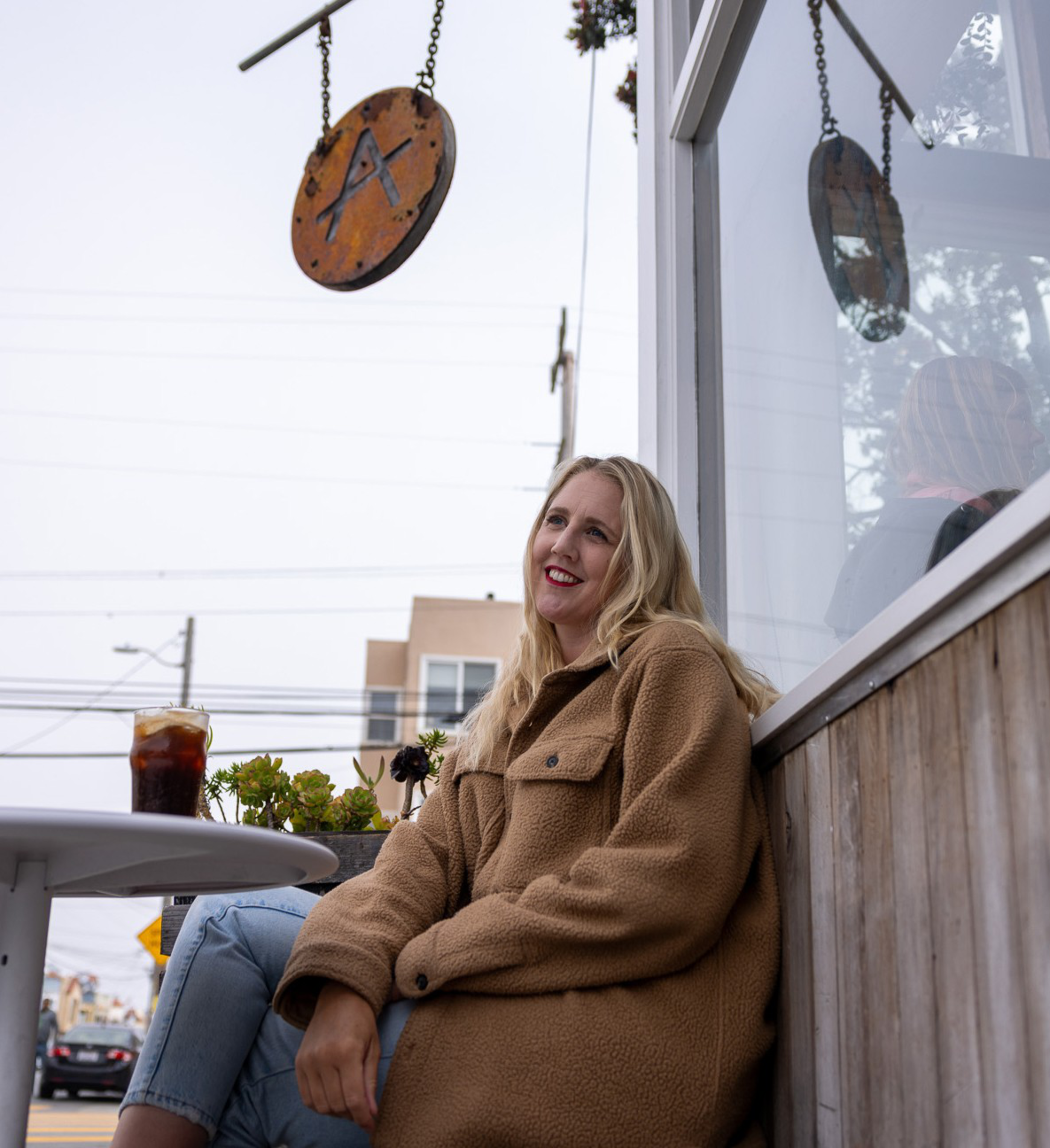 A woman in a tan coat sits outside a café, smiling. A glass of dark iced beverage is on the table, and a round café sign with an &quot;A&quot; hangs above her.