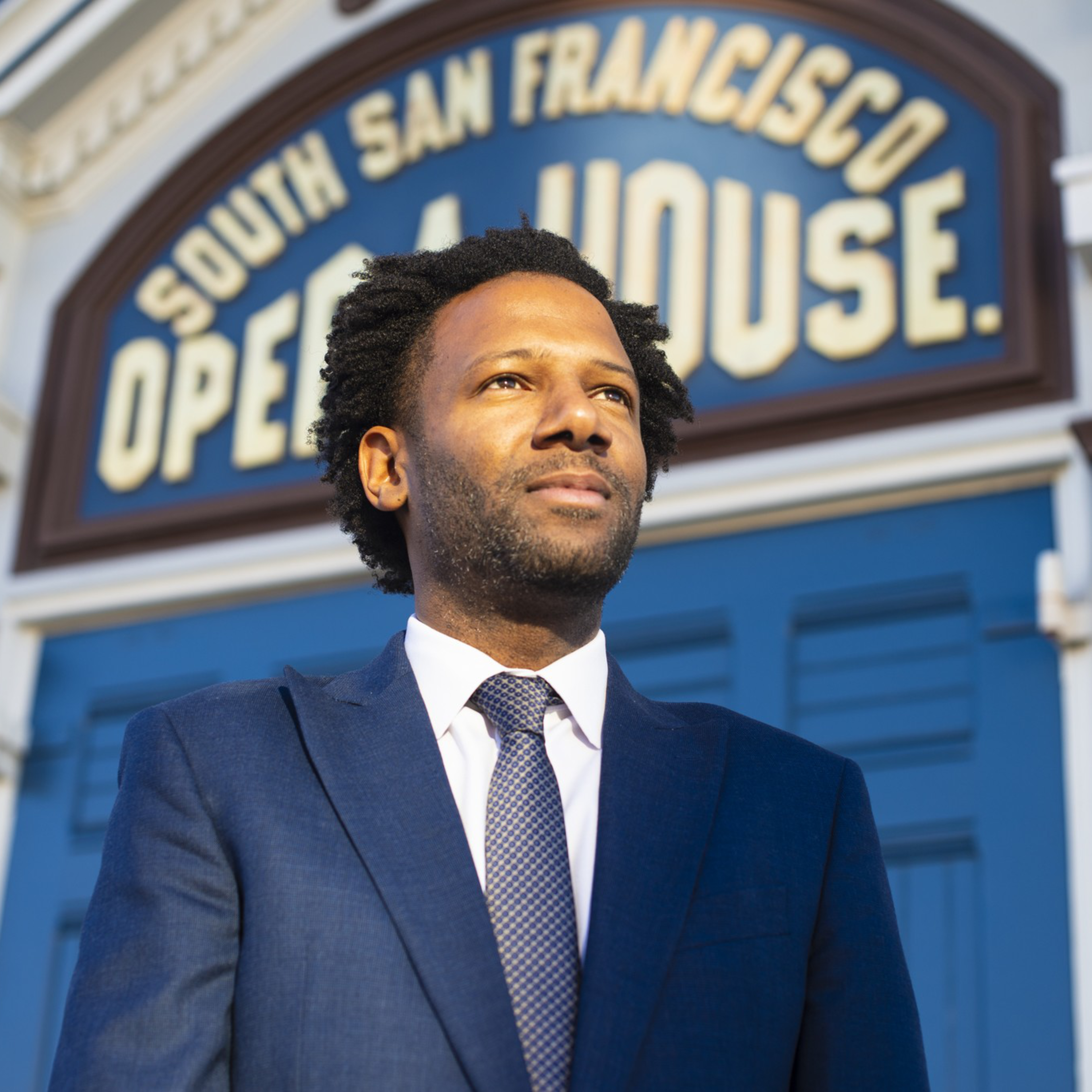 A man in a blue suit and tie stands confidently in front of a building with a sign that reads "South San Francisco Opera House."