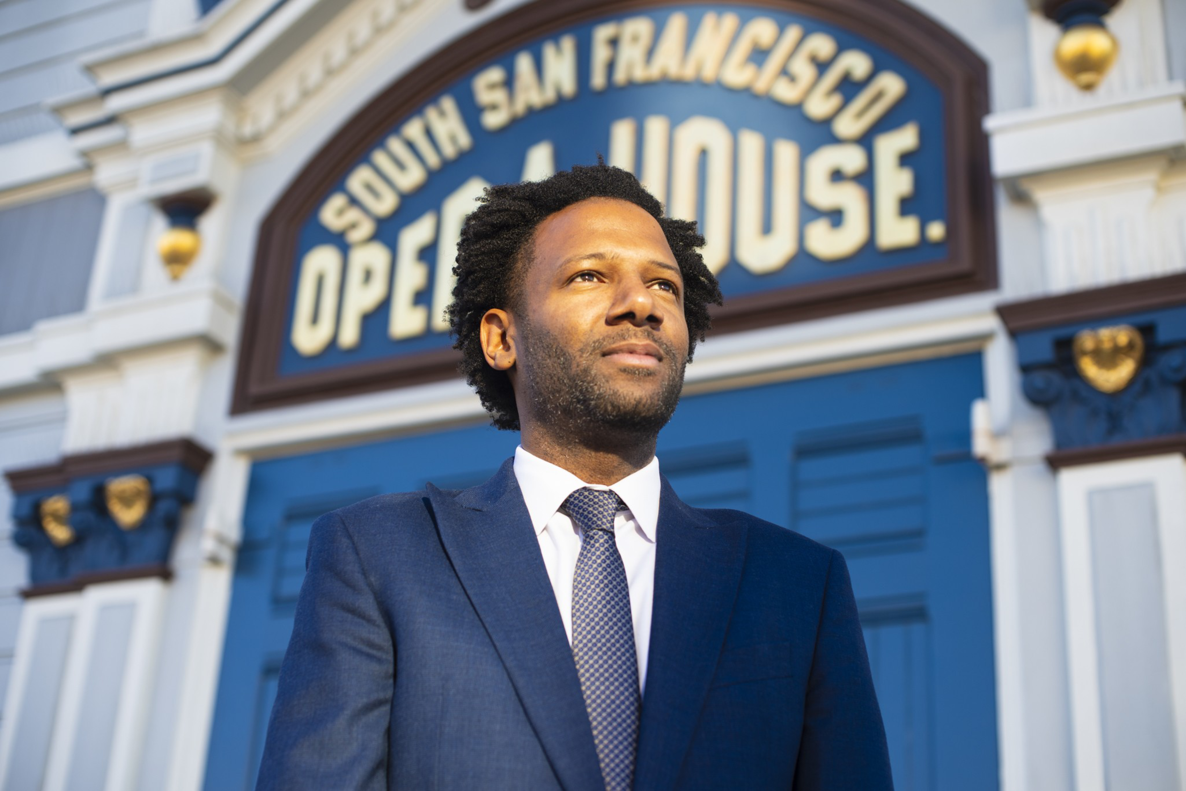 A man in a blue suit and tie stands confidently in front of a building with a sign that reads "South San Francisco Opera House."