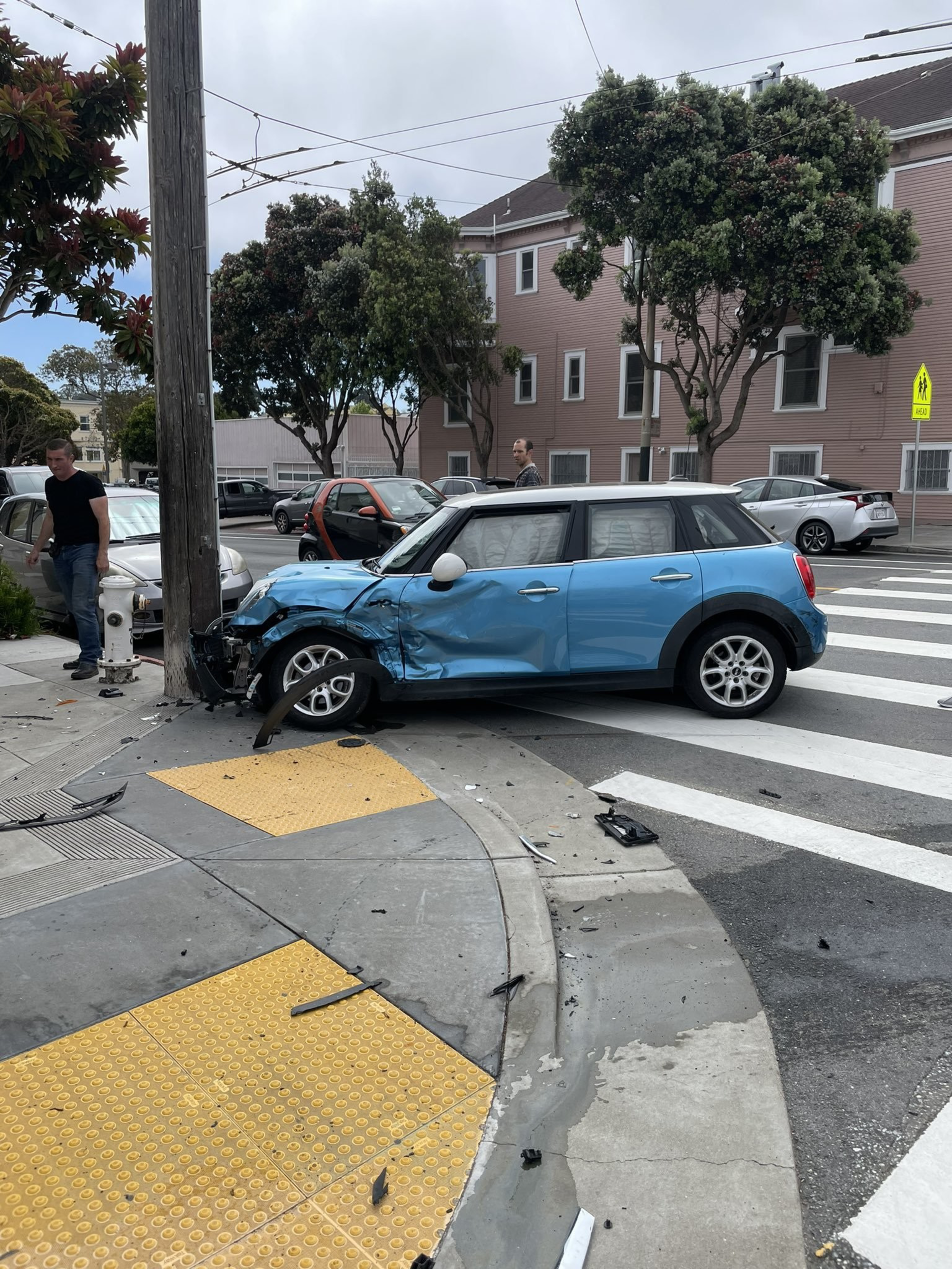 A blue car with severe front damage is on a street corner near a traffic pole; debris is scattered on the road. Two men stand nearby observing.