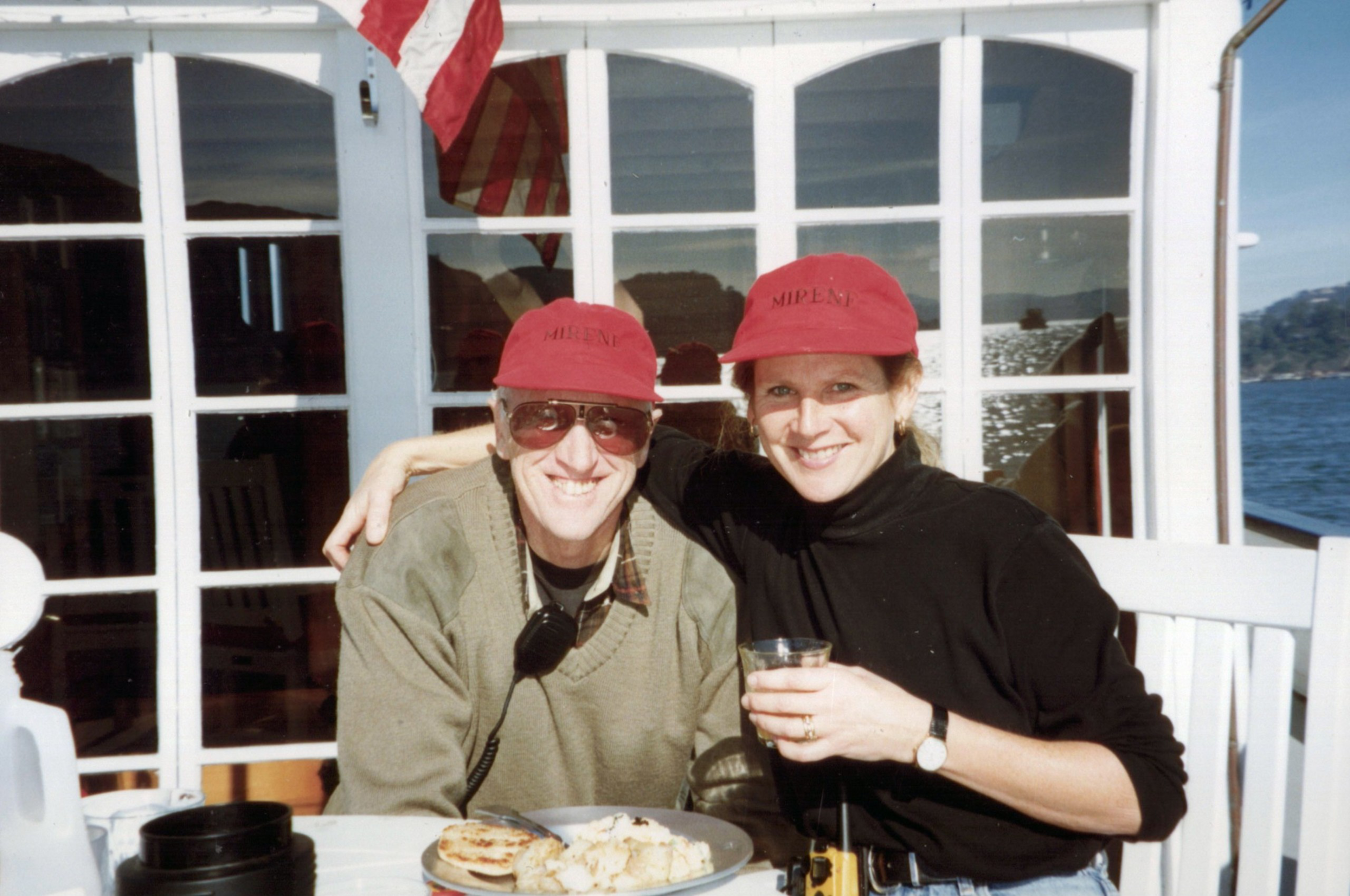 Two people wearing matching red caps are sitting close, smiling; one holds a drink, while a plate with food sits in front of them on a white table.
