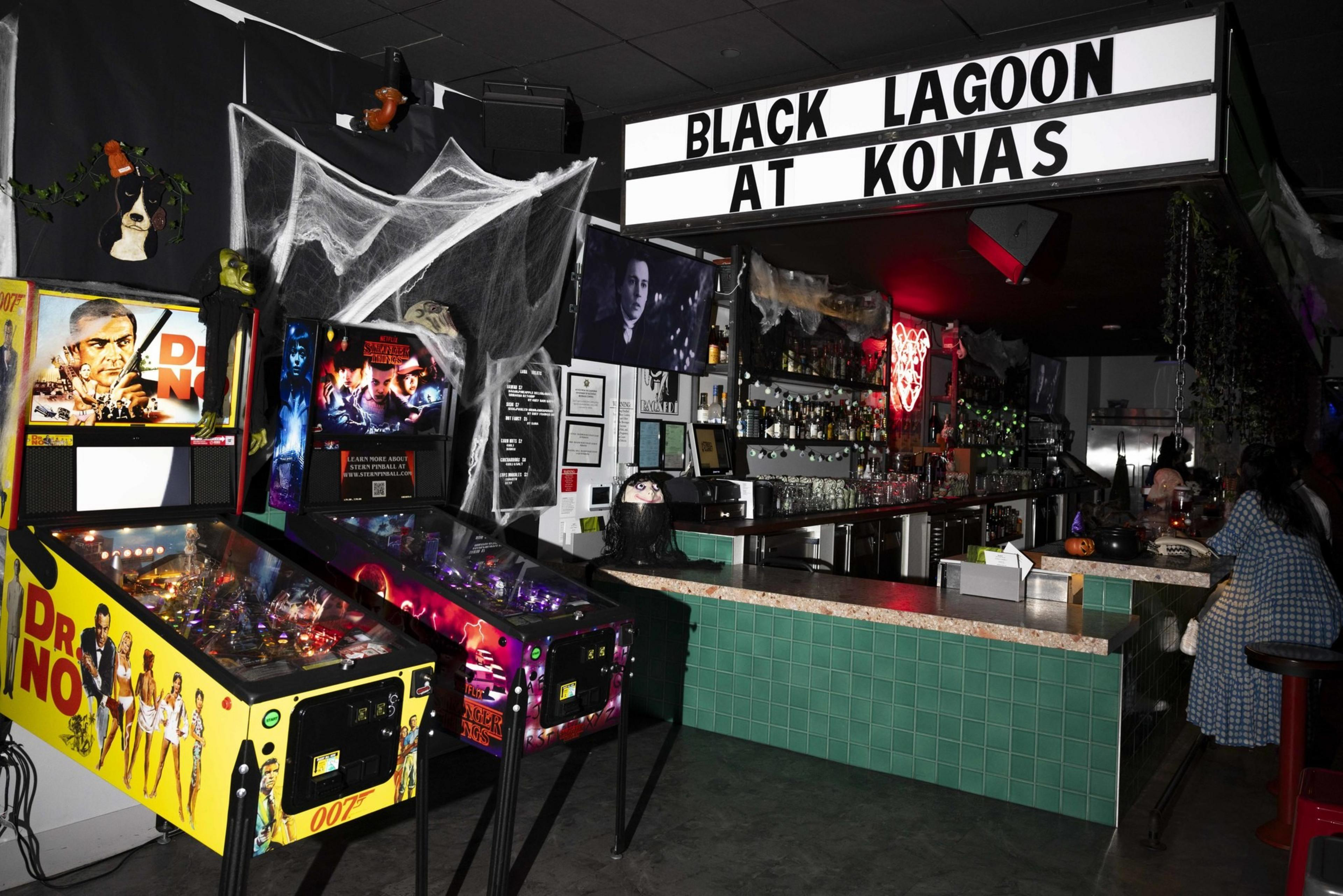 The image shows a bar with green tiles, two pinball machines, spooky decorations, and a sign saying &quot;BLACK LAGOON AT KONAS.&quot; There's a person at the bar.