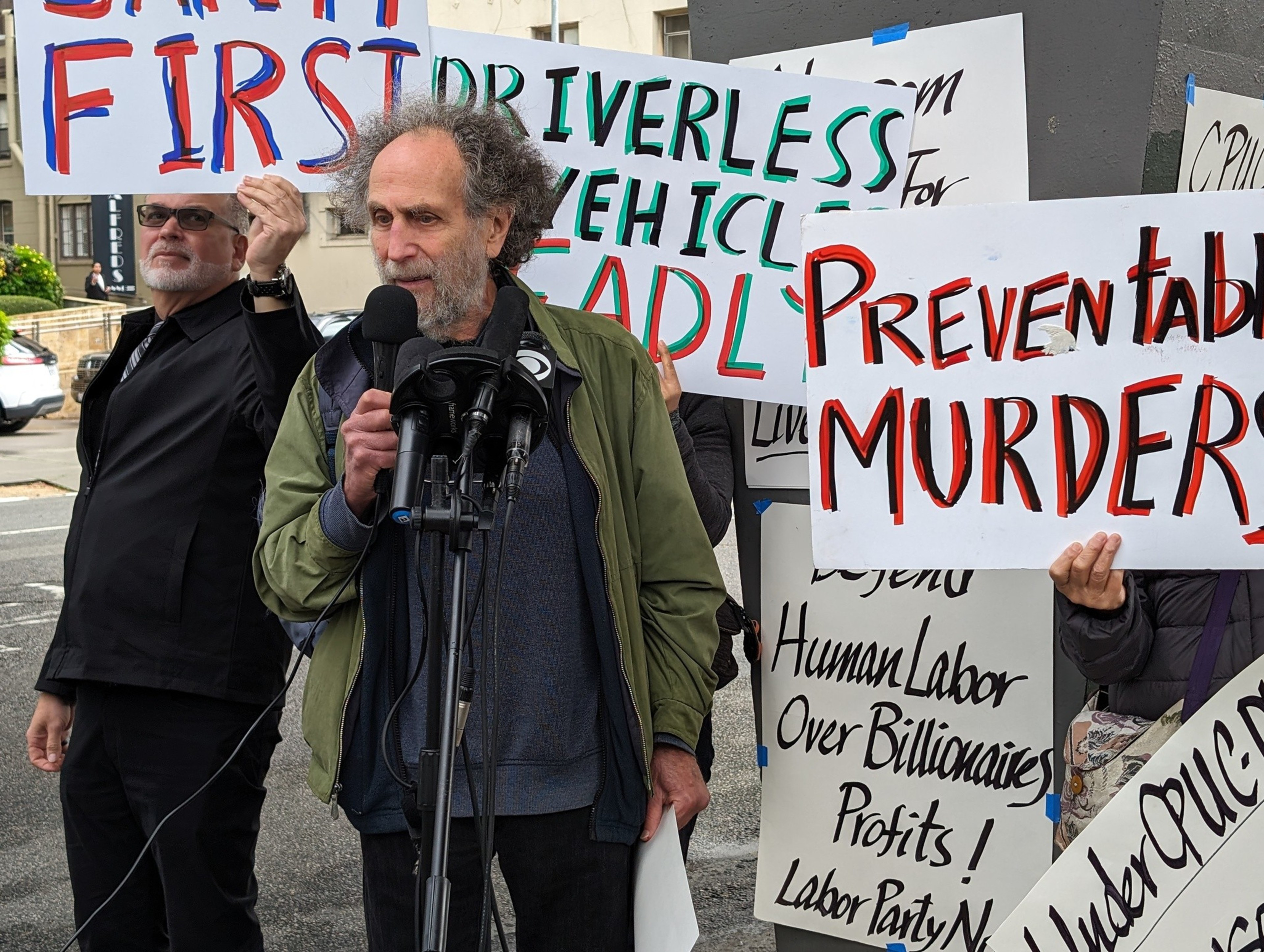 A man holds a posterboard sign and stands next to another man speaking into a stand for multiple microphones.