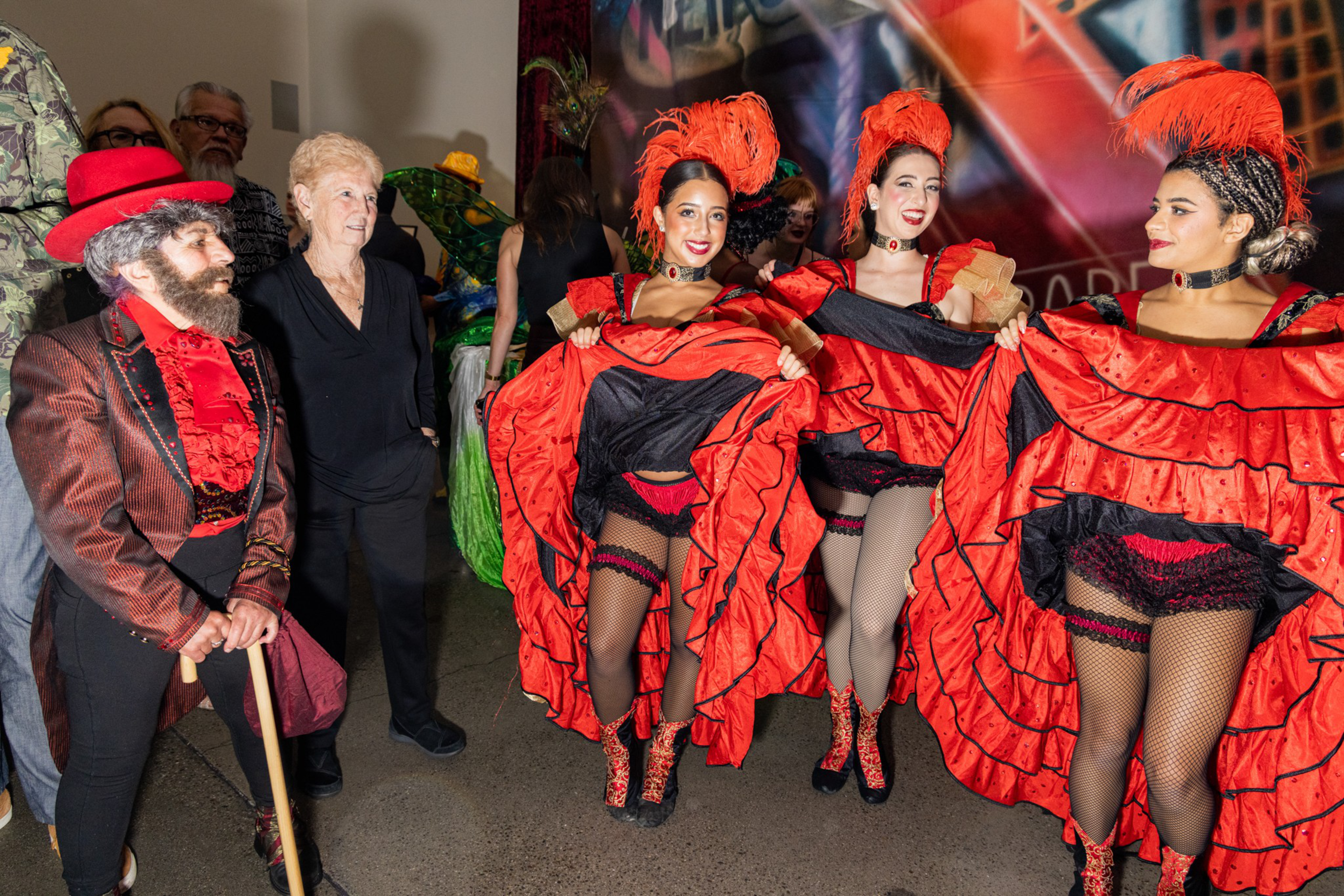 A group of people dressed in elaborate costumes, including a bearded man in red and black and three women in red can-can dresses, with an older woman in black nearby.