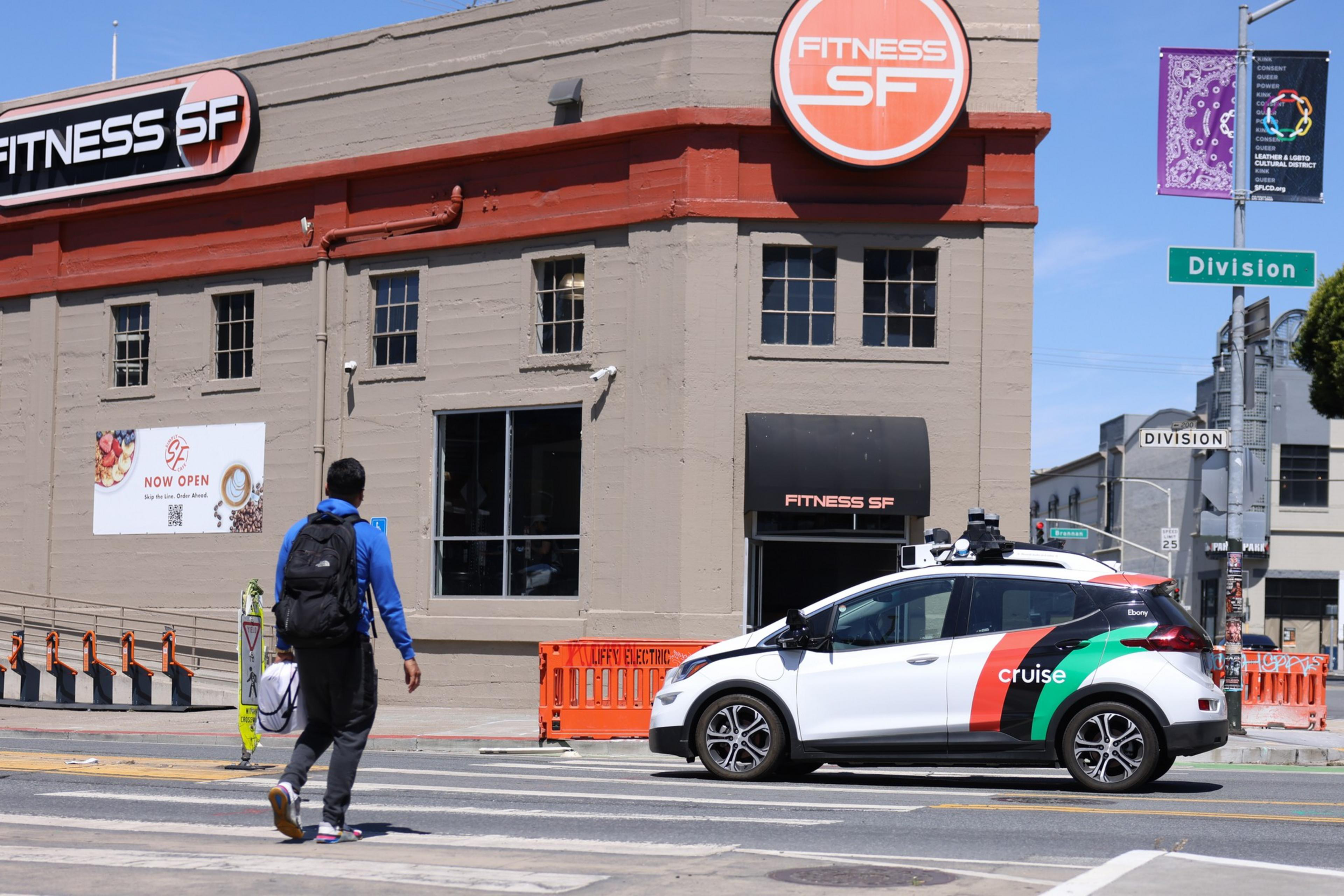 A car without a driver is seen in the road by a building as a man uses a crosswalk nearby