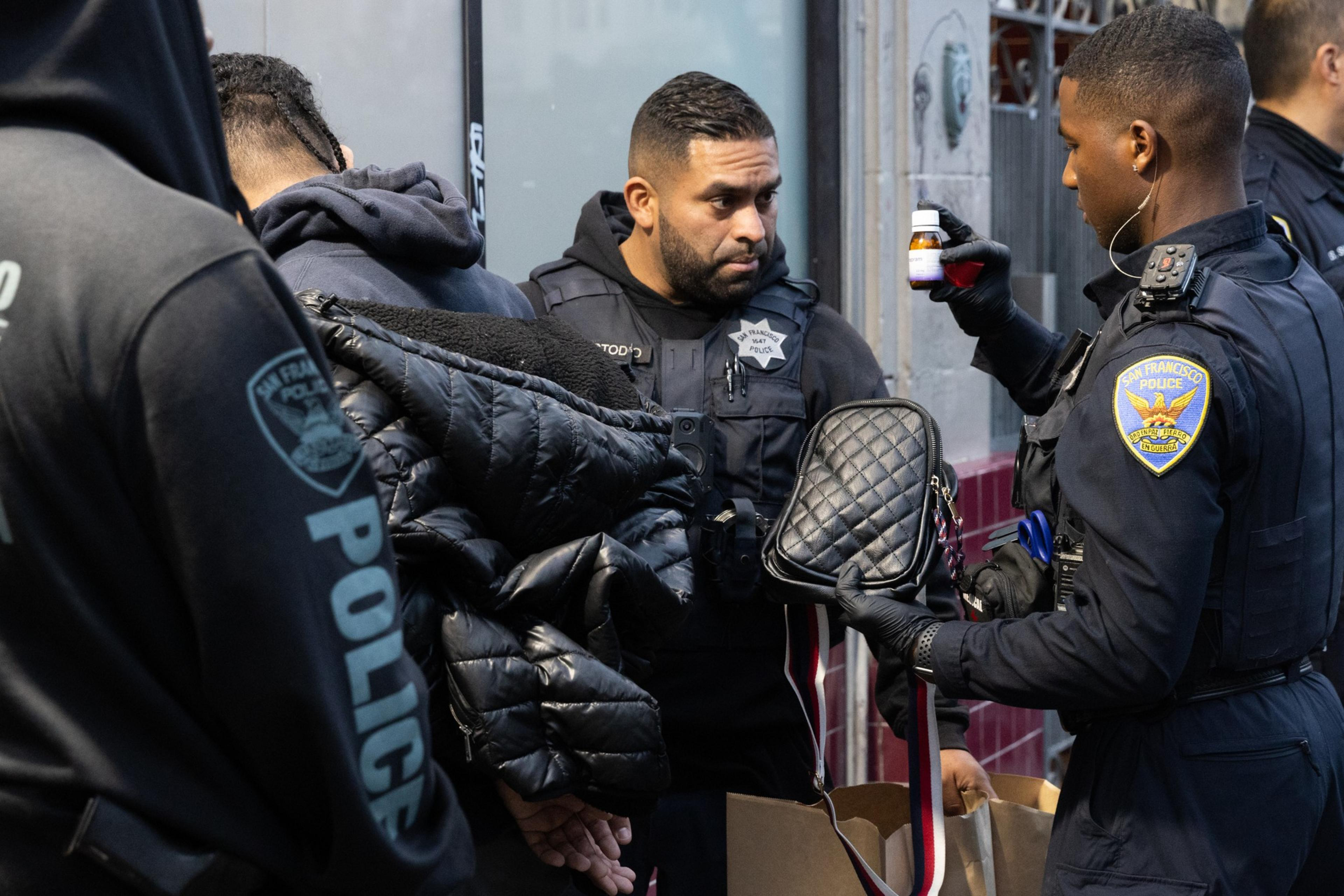 Two police officers in uniform are examining a small bottle and a quilted bag. Another person is visible wearing a hooded jacket.