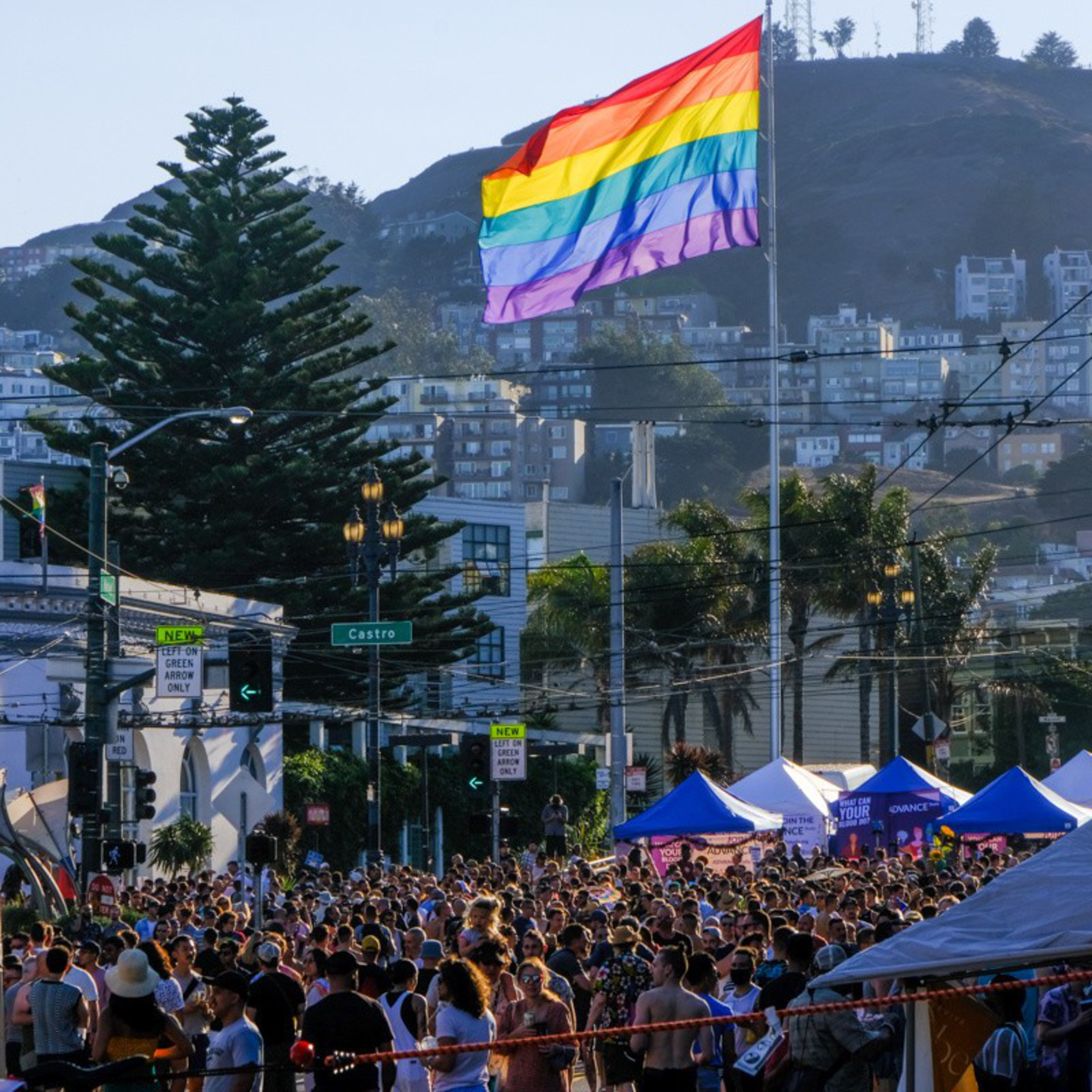A large crowd gathers under a rainbow pride flag in a cityscape with hills, trees, and buildings visible in the background on a sunny day.