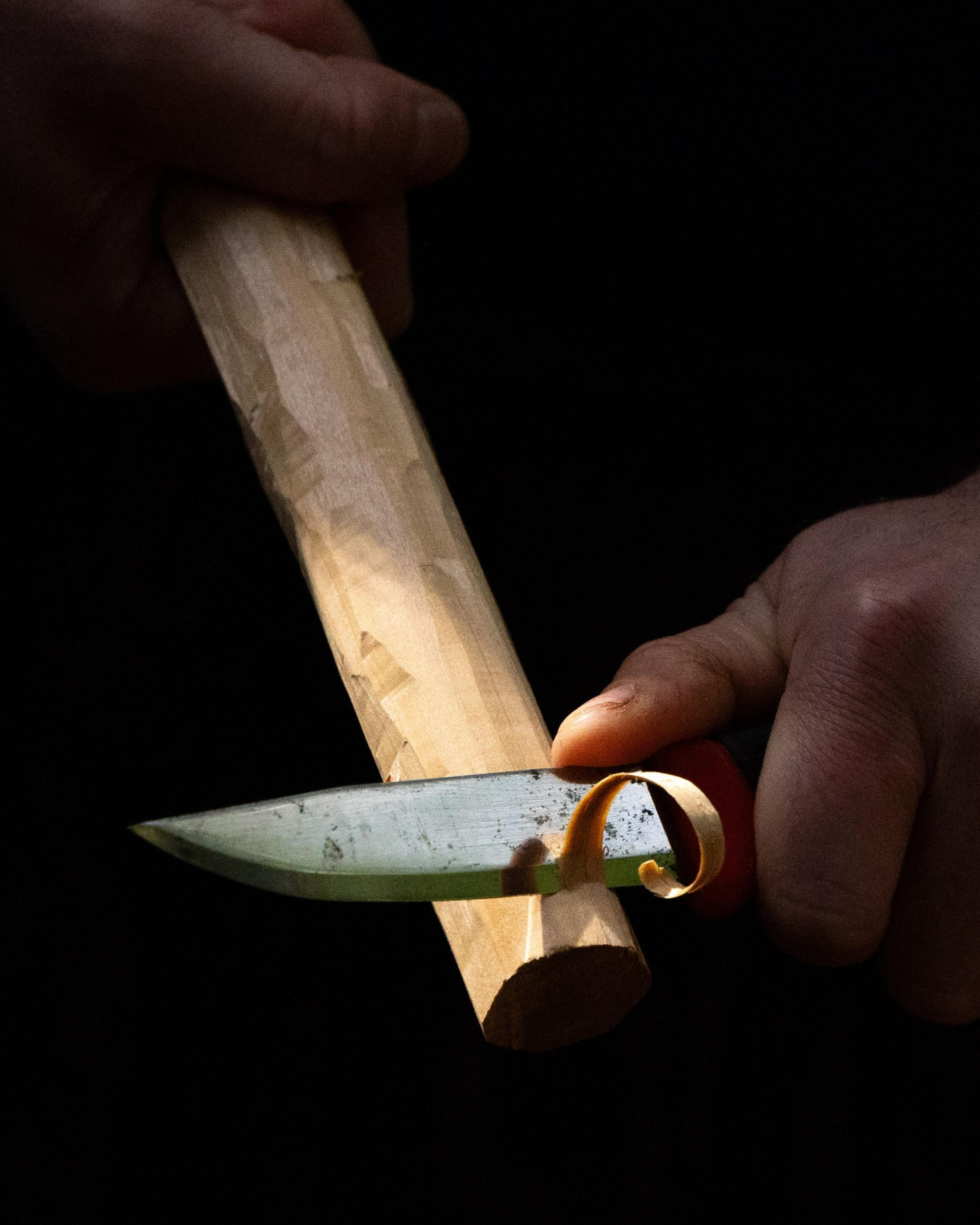 A person is carving a piece of wood with a knife. The person holds the wood steady while slicing a thin, curled shaving from it with the blade.
