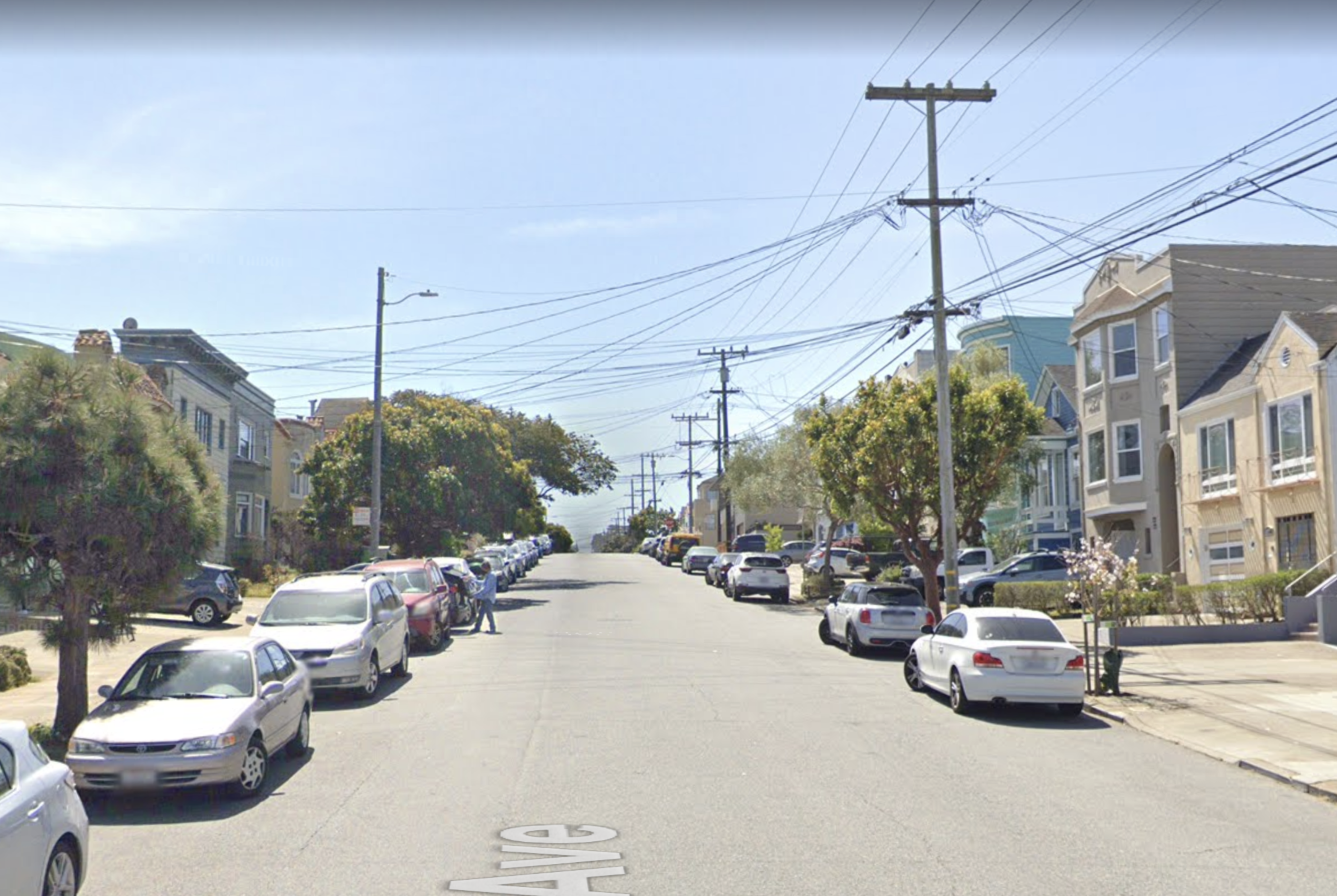 A quiet residential street lined with parked cars on both sides, trees, utility poles with overhead wires, and houses under a clear blue sky.