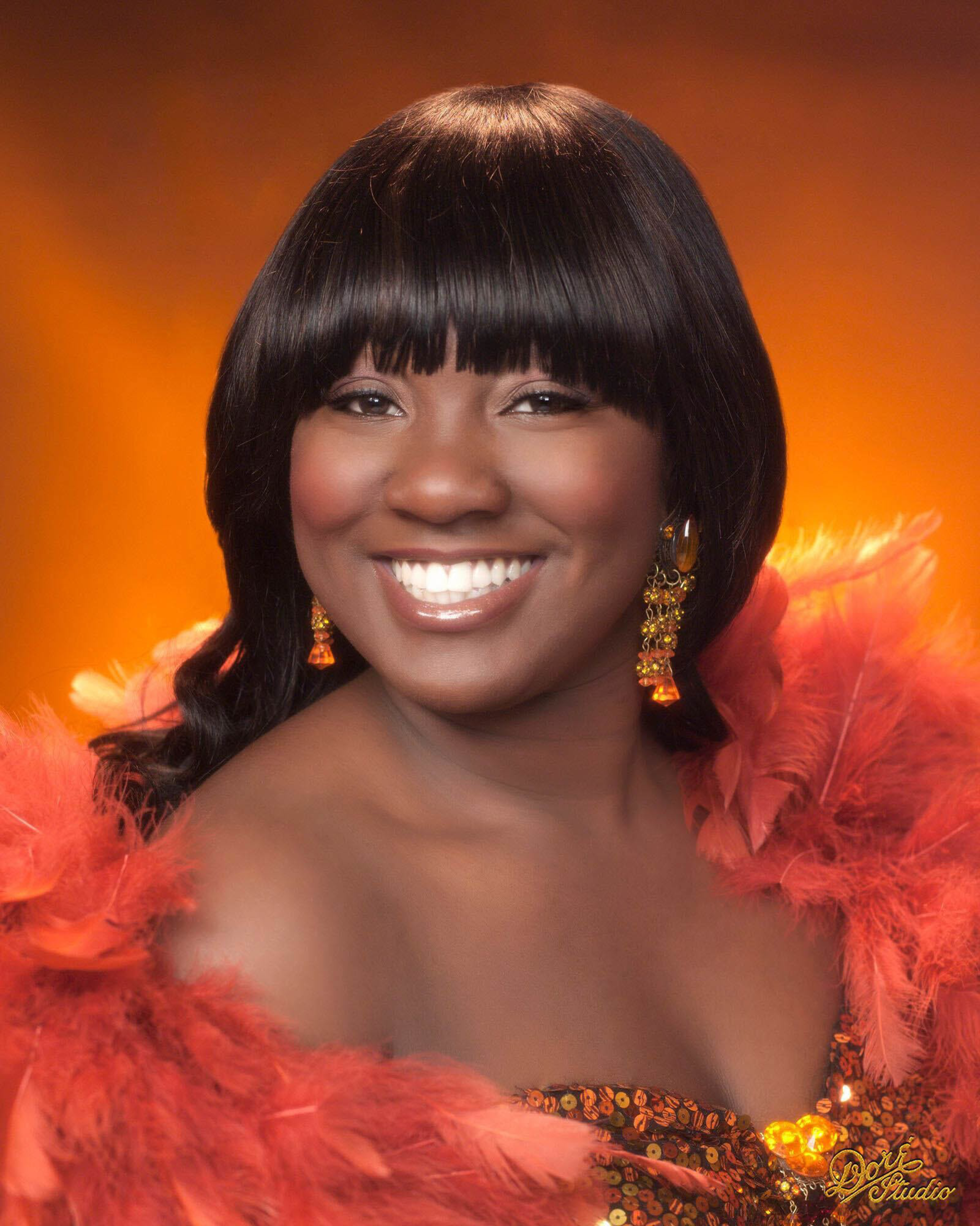 A smiling woman with straight black hair and bangs wears orange feathered attire and matching dangling earrings, set against a warm orange background.